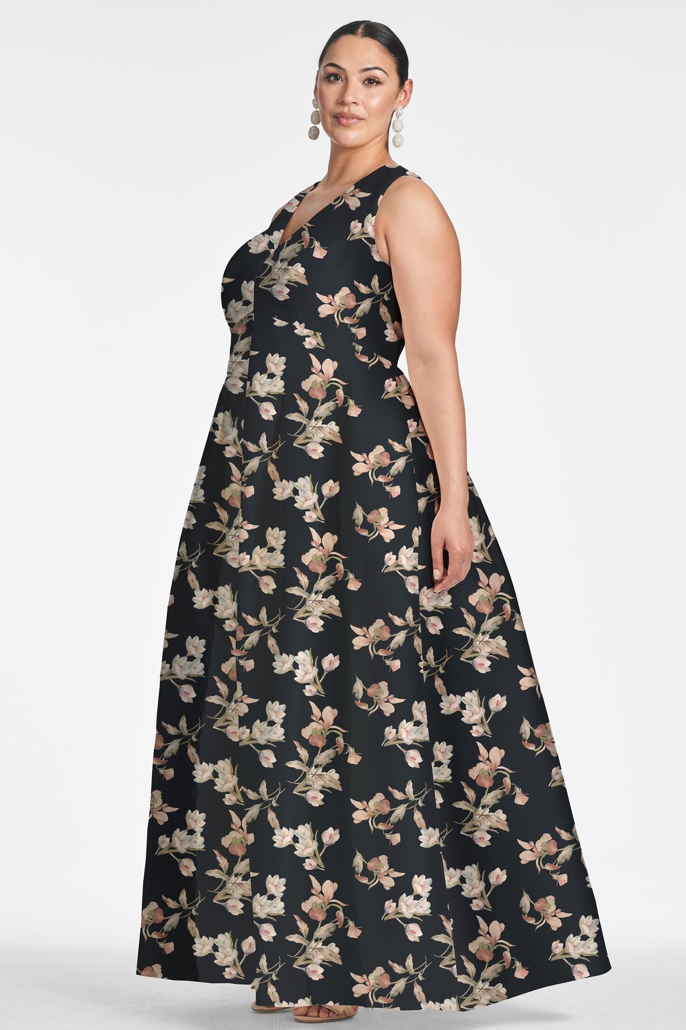 A woman stands confidently, modeling the Sachin & Babi Brooke Gown - Tonal Petal. This sleeveless black V-neck gown is adorned with an intricate floral pattern and features side pockets and box pleats. She complements the look with dangling earrings and her hair pulled back. The plain white background draws attention to her stylish dress from Sachin & Babi.