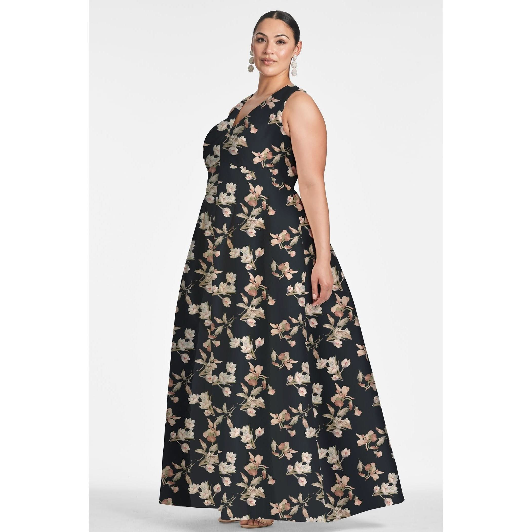 A woman stands confidently, modeling the Sachin & Babi Brooke Gown - Tonal Petal. This sleeveless black V-neck gown is adorned with an intricate floral pattern and features side pockets and box pleats. She complements the look with dangling earrings and her hair pulled back. The plain white background draws attention to her stylish dress from Sachin & Babi.