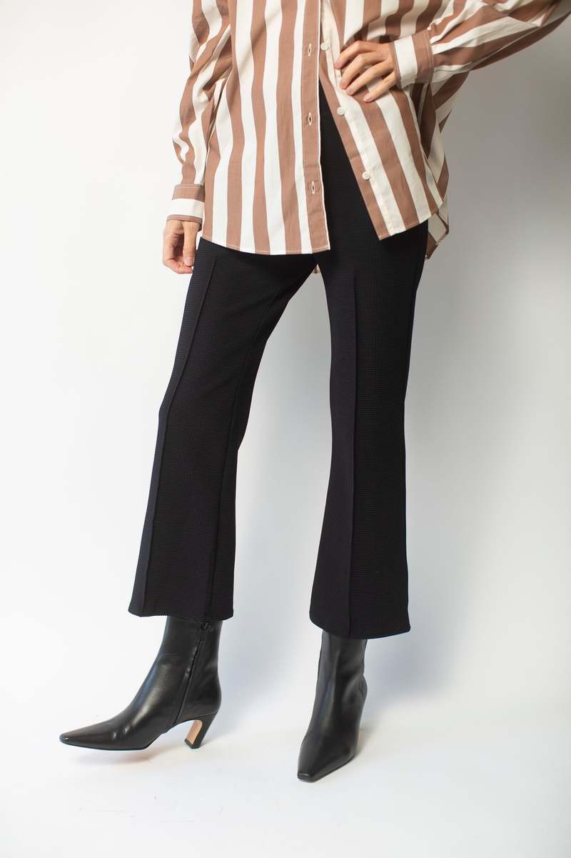 A person is standing against a white background wearing a brown and white striped shirt paired with Donni The Box Weave Stretch Kick Flare pants by Donni and black ankle boots with low heels. Only their legs and torso are visible.