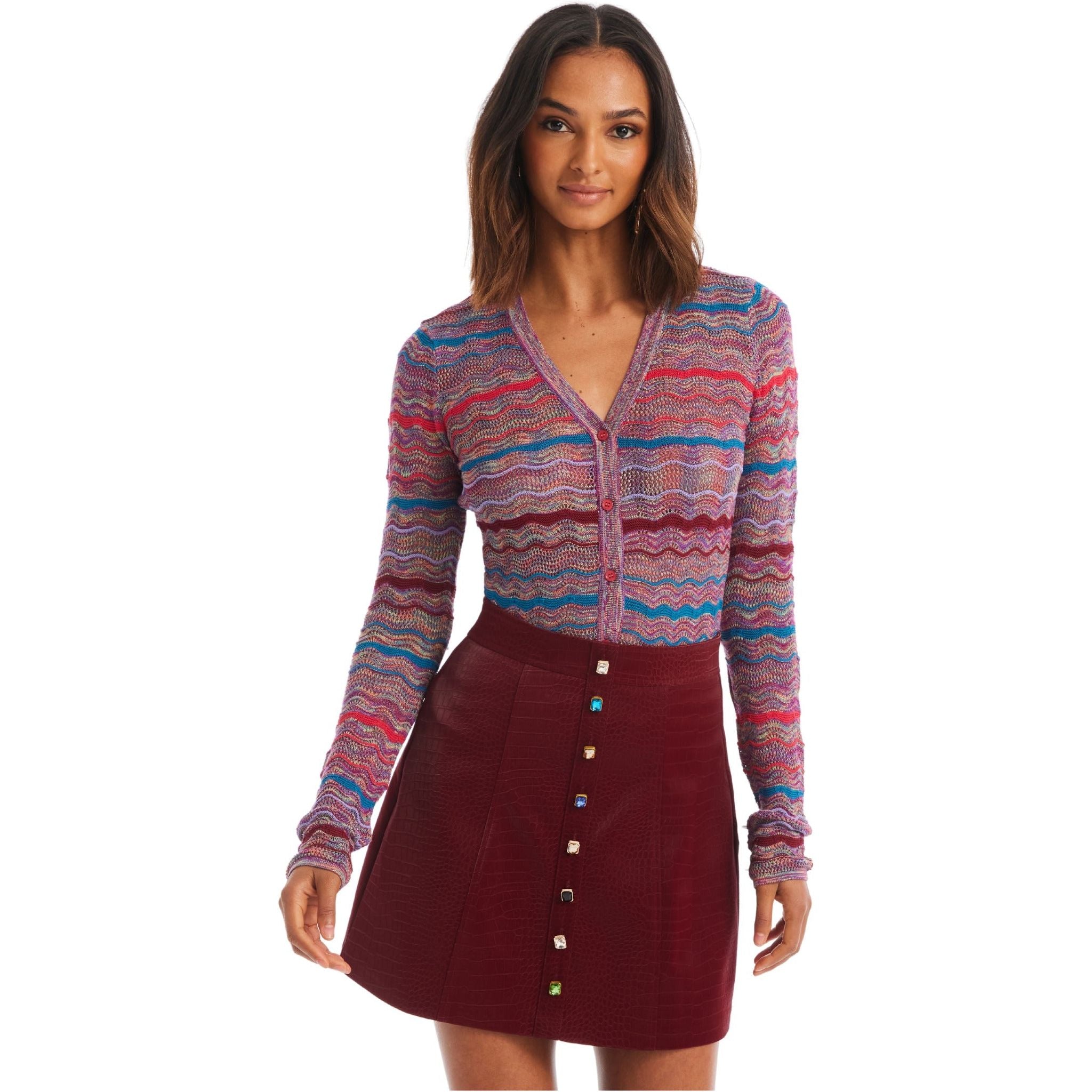 A woman stands facing the camera, wearing the Allison NY COLLEEN CARDIGAN by Allison, characterized by its long sleeves and multicolored wavy pattern in an oversized fit. She pairs it with a high-waisted red buttoned skirt. Her long, wavy hair cascades as she shows a slight smile on her face.