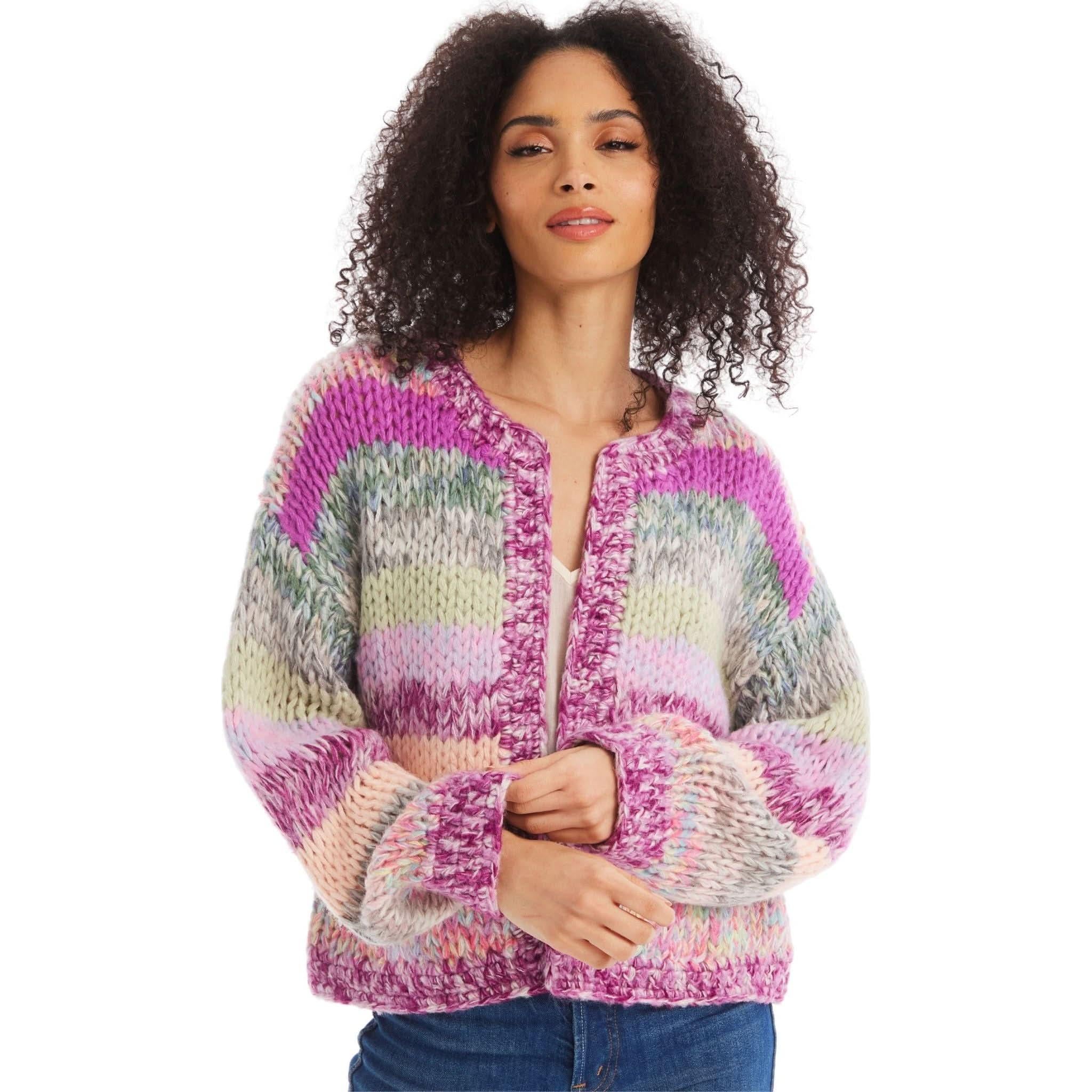 A woman with curly hair is wearing an Allison NY Cropped Jodie Cardigan by Allison, featuring an oversized, chunky-knit design in shades of pink, purple, green, and beige. The cropped-length cardigan has a textured, cozy look and is dry clean only. She pairs it with a white top and blue jeans as she stands against a plain white background, smiling gently.