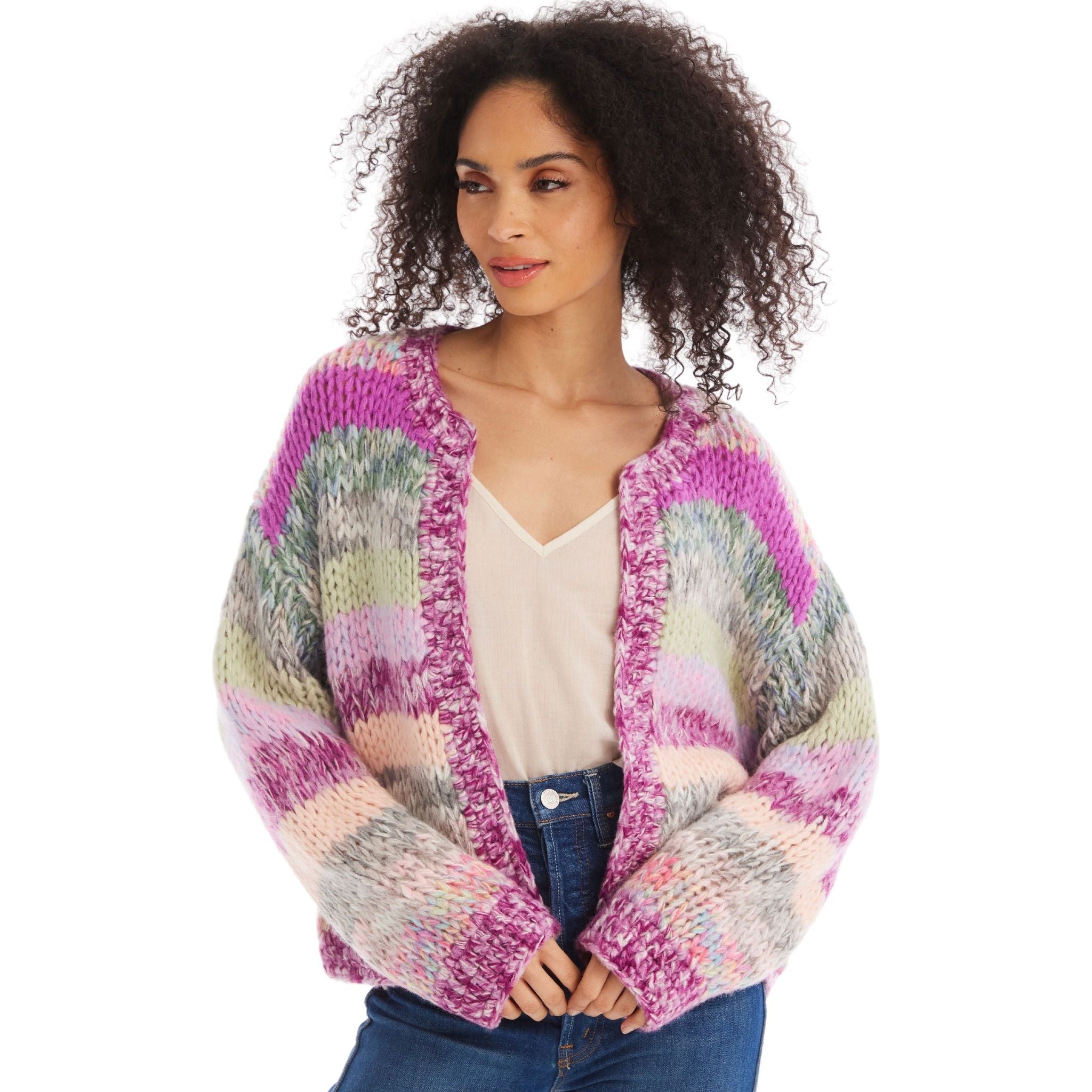 A person with curly hair wears the Allison NY Cropped Jodie Cardigan from Allison, featuring vibrant pink, purple, green, and gray stripes over a white top. The chunky knit cardigan's oversized fit gives them a cozy look as they gaze to one side with hands resting at their waist. They are also wearing blue jeans.