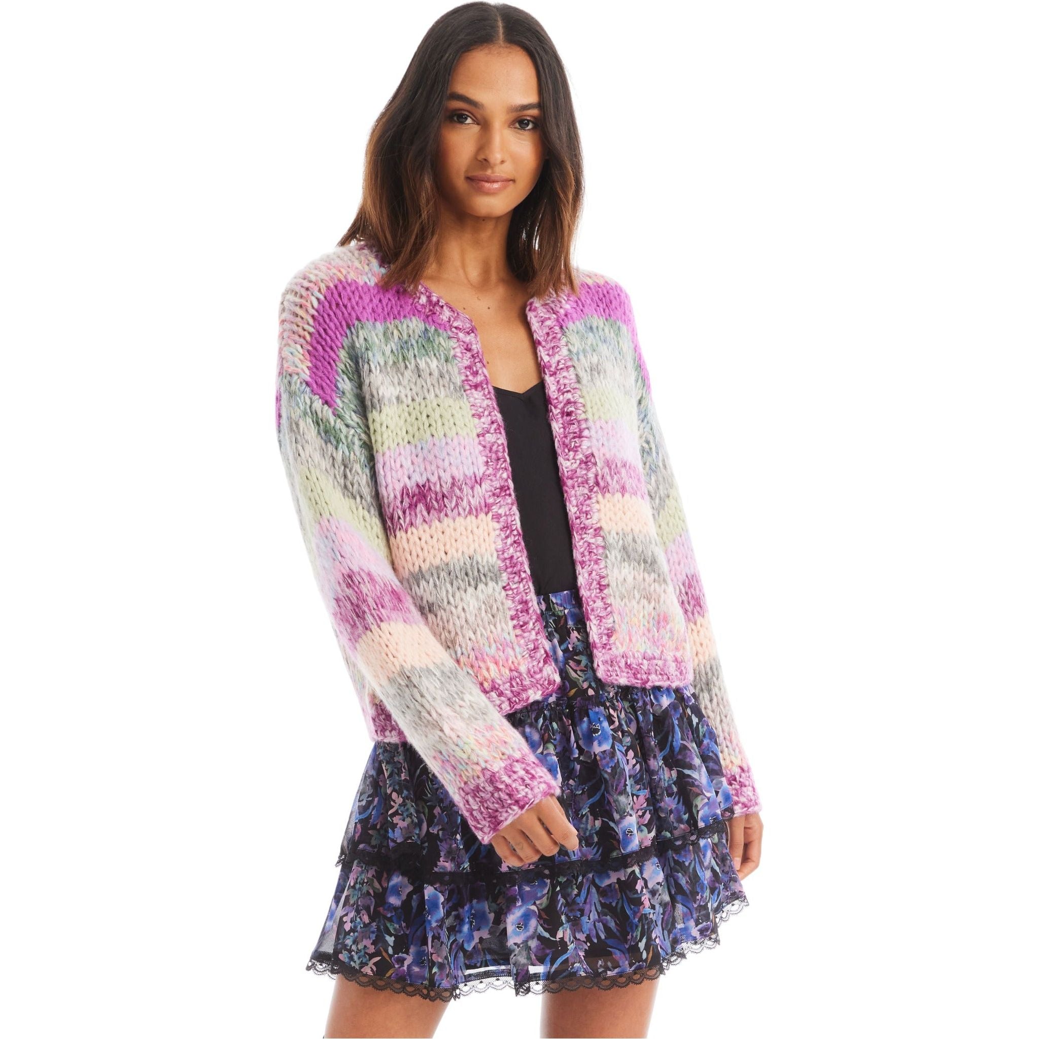 A woman with shoulder-length hair wears the Allison NY Cropped Jodie Cardigan, a vibrant chunky knit cardigan from Allison in shades of pink, green, and purple over a black top. She pairs it with a layered, ruffled skirt featuring a blue and purple floral pattern. This stylish outfit is dry clean only. She stands against a white background.