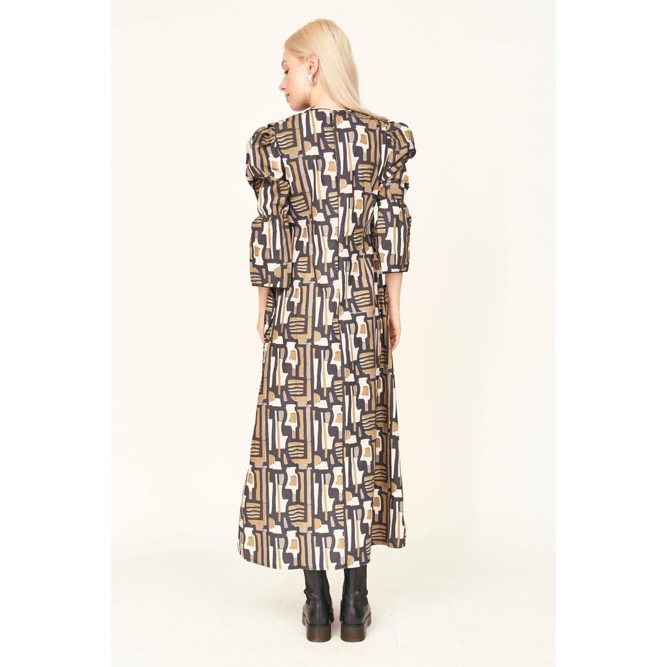 A person with long blond hair is wearing the Mirth Cassis Dress in Olive Labyrinth, a cotton poplin piece featuring a design of geometric shapes in black, white, and gold. They are standing and facing away from the camera while paired with black boots.