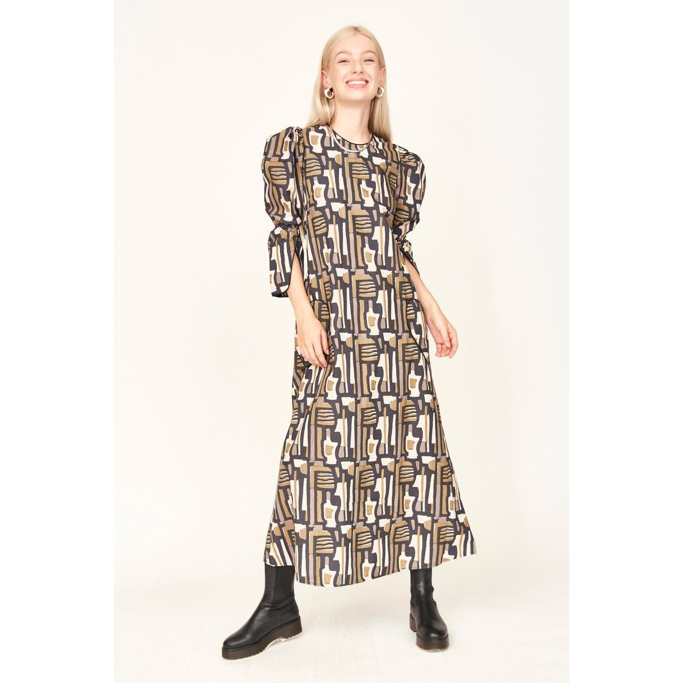 A person smiling, dressed in the Mirth Cassis Dress in Olive Labyrinth, an ankle-length hand block printed dress with puffed sleeves, paired with black boots. Their long blonde hair cascades down as they stand confidently against a plain background.