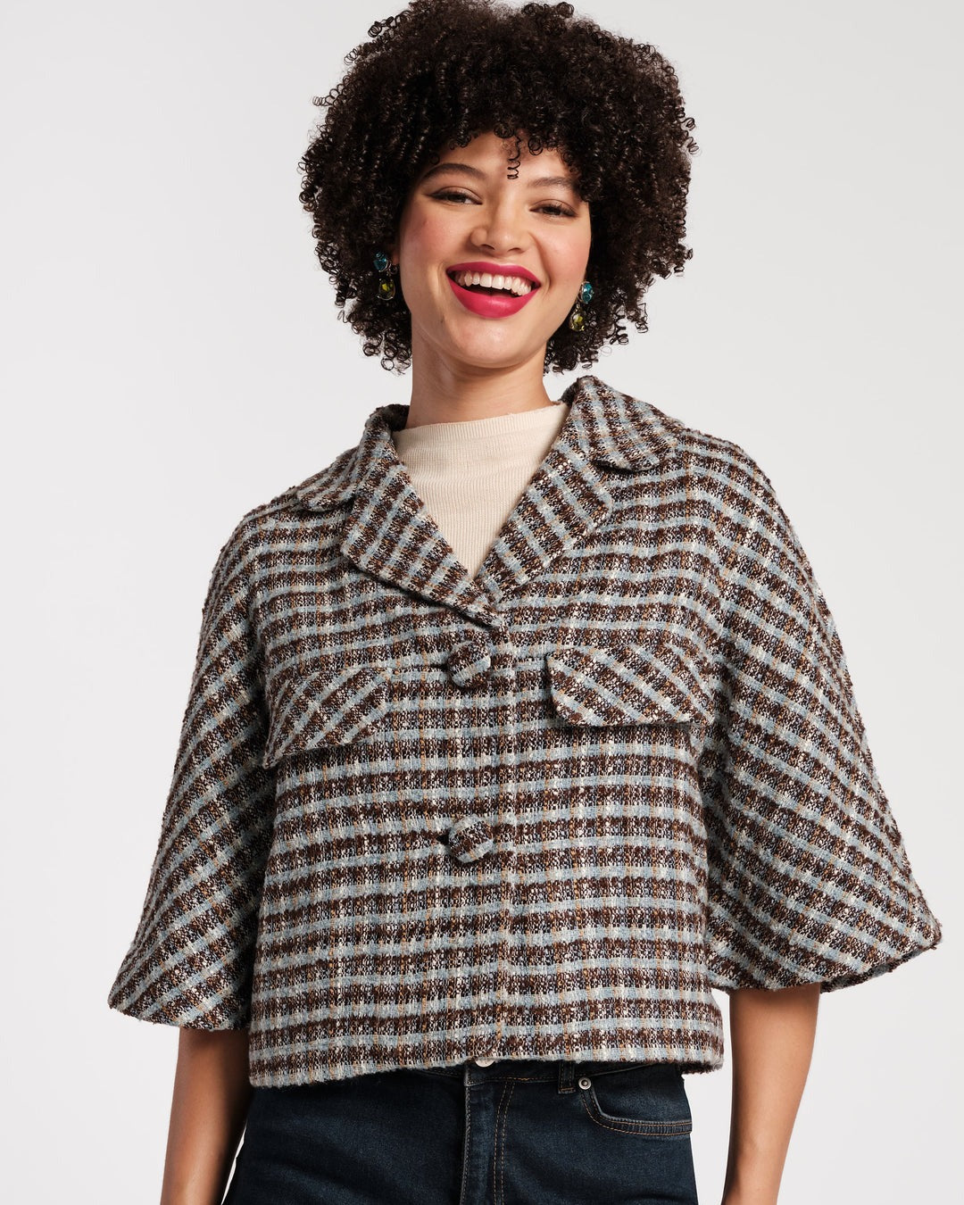 A person stands against a plain background wearing a Frances Valentine Day Jacket in Wool Park Avenue Plaid Blue Brown, a light beige top, and brown pants. They are holding a pink handbag and have on gold shoes. Their curly hair is styled naturally, and they are accessorized with bracelets.