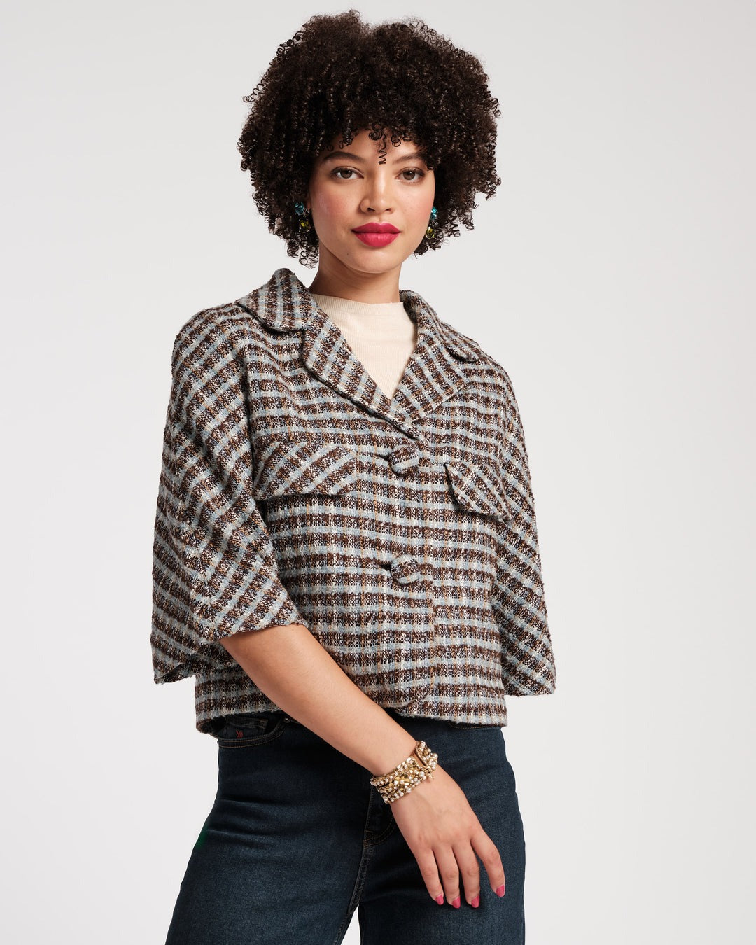 A person stands against a plain background wearing a Frances Valentine Day Jacket in Wool Park Avenue Plaid Blue Brown, a light beige top, and brown pants. They are holding a pink handbag and have on gold shoes. Their curly hair is styled naturally, and they are accessorized with bracelets.