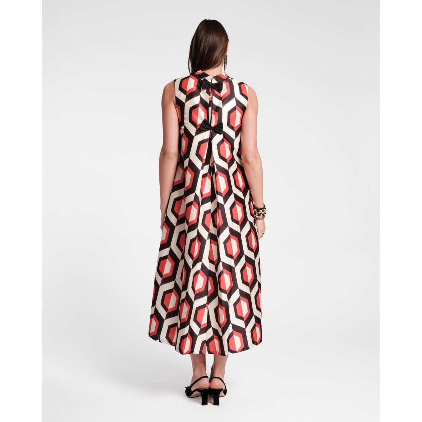 A woman stands facing away, displaying the Frances Valentine Diplomat Maxi Dress in an elegant empire print jacquard featuring oyster and red geometric patterns. Her long dark hair flows down as she pairs the dress with black sandals and bracelets, all set against a simple white backdrop.