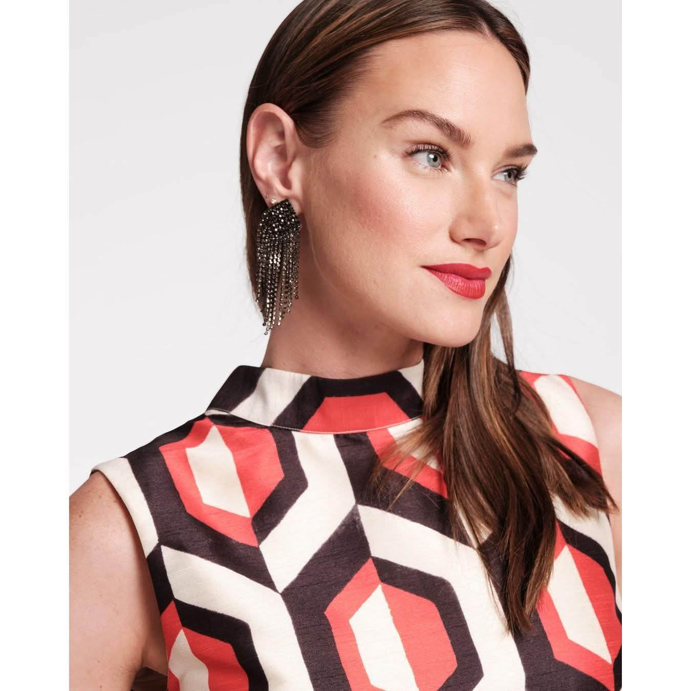 A woman with long brown hair is wearing the Frances Valentine Diplomat Maxi Dress featuring an Empire Print Jacquard pattern in oyster and red. She has large dangling earrings and is looking to the left with a slight smile, set against a plain white background.