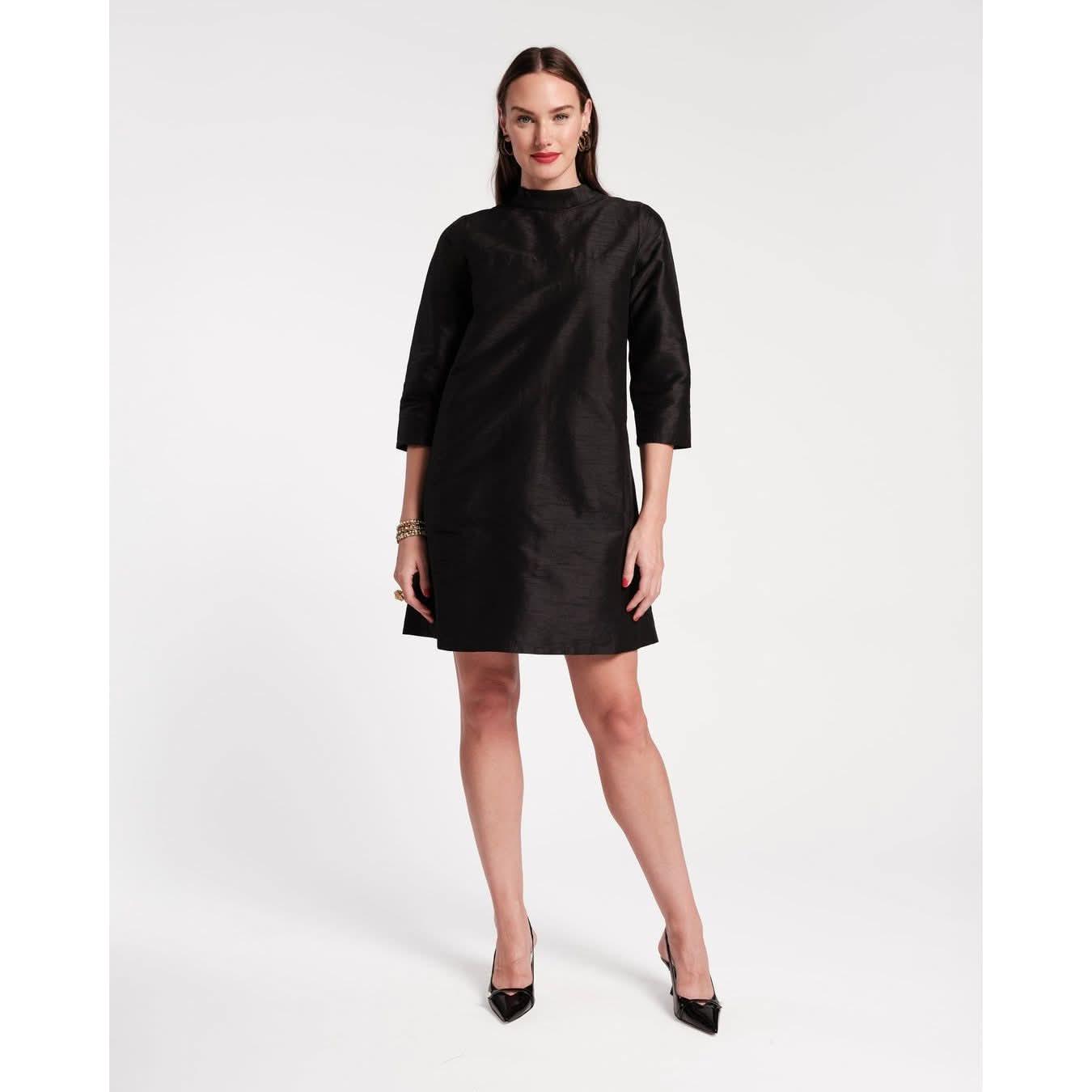 A person poses in the Frances Valentine Diplomat Mini Dress Black, showcasing its simple and elegant style with three-quarter sleeves. They stand against a plain white background, wearing black pointed heels and keeping their hands relaxed by their sides.