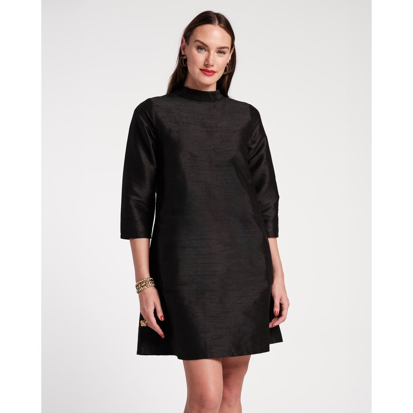 A person stands confidently in a sleek, black Frances Valentine Diplomat Mini Dress with three-quarter sleeves. Their long, straight hair complements their hoop earrings and bracelets. The backdrop is a simple white, highlighting the elegance of their ensemble.