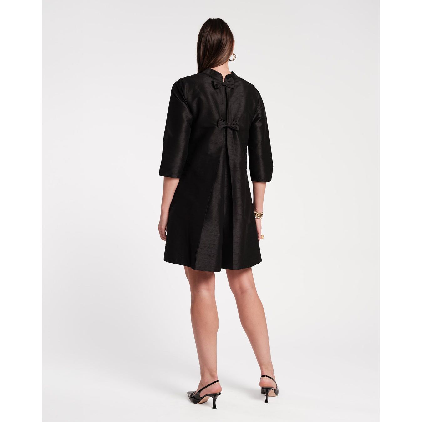 A woman stands facing away in the Frances Valentine Diplomat Mini Dress in black, crafted from lustrous dupioni and adorned with a bow detail at the back. The dress features elegant three-quarter sleeves, complemented by her stylish black heels against a plain white backdrop.