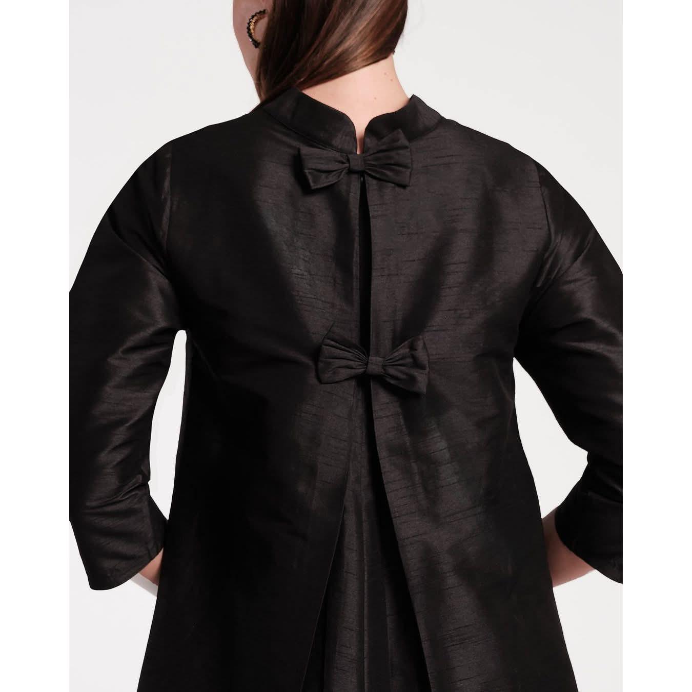 A person wearing the Frances Valentine Diplomat Mini Dress Black, crafted from solid Dupioni fabric, features two decorative bows along the back seam. The design elegantly splits open to reveal a layered, pleated fabric underneath. This dress is characterized by a high collar and long sleeves for added sophistication.