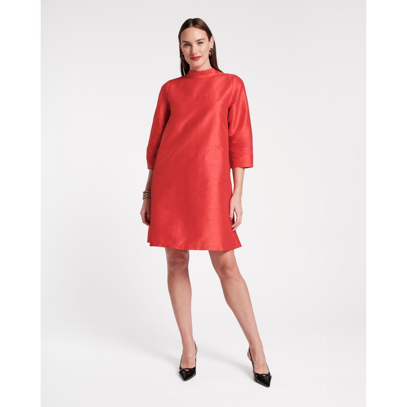 A person confidently stands in the Frances Valentine Diplomat Mini Dress Red, featuring a knee-length design with a high neckline, three-quarter sleeves, and subtle pleats. They sport black high heels and accessorize with a gold bracelet against a plain white background.