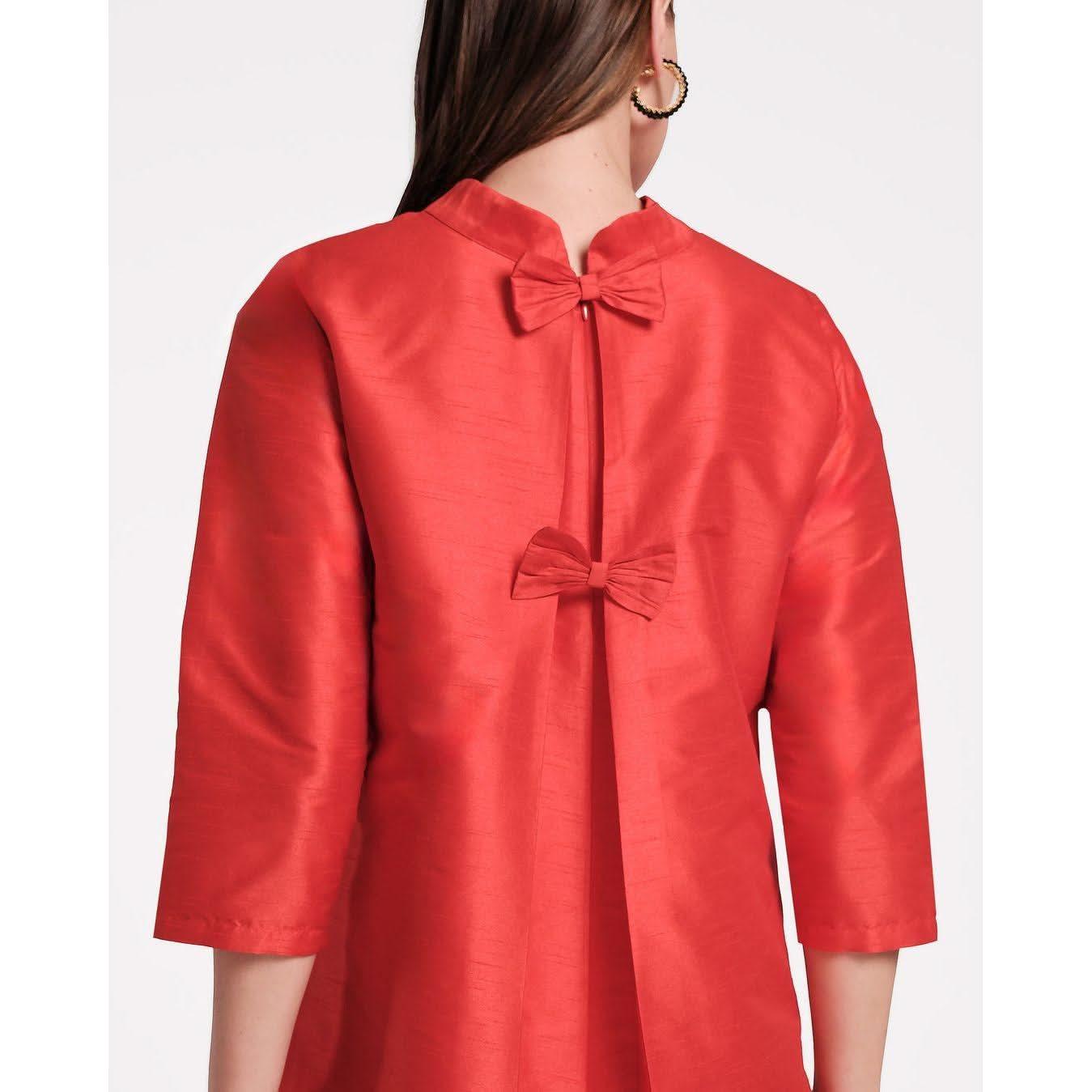 A person with long hair is shown from behind, wearing the Frances Valentine Diplomat Mini Dress in red, which features a shiny finish, a mock neckline, two decorative bows on the back, and elbow-length sleeves.