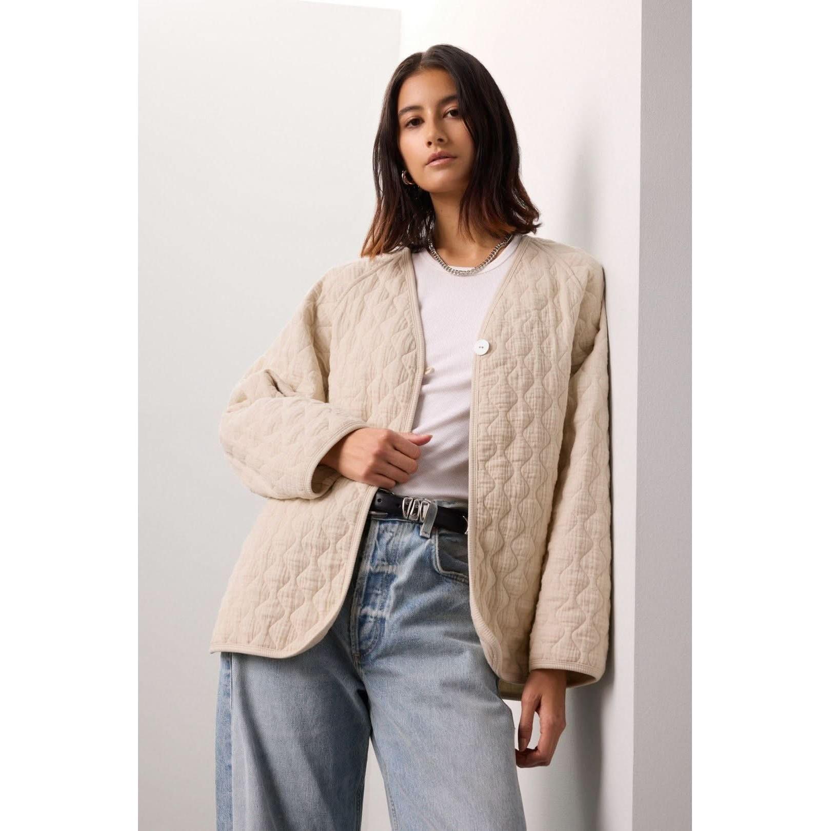 A person wearing the Donni The Quilted Jacket Creme over a white shirt and light blue jeans stands against a white wall. They have shoulder-length dark hair, accessorized with a necklace made of organic cotton. The soft and even lighting highlights the delicate textures in their attire.