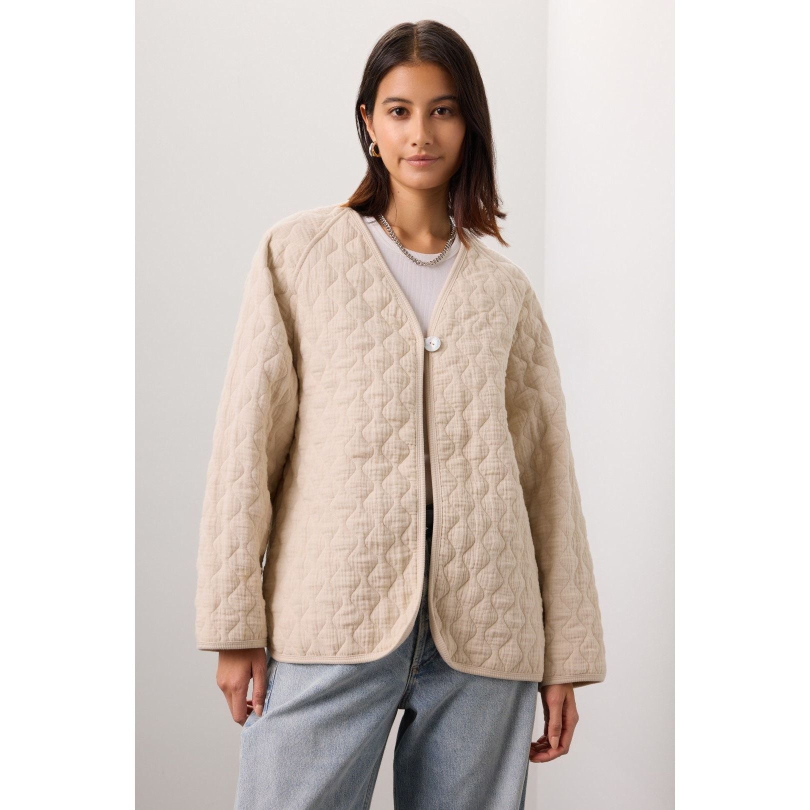 A person stands against a neutral background wearing Donni's The Quilted Jacket in Creme, featuring a button closure over an organic cotton shirt and light blue jeans. They have shoulder-length hair and a subtle smile.