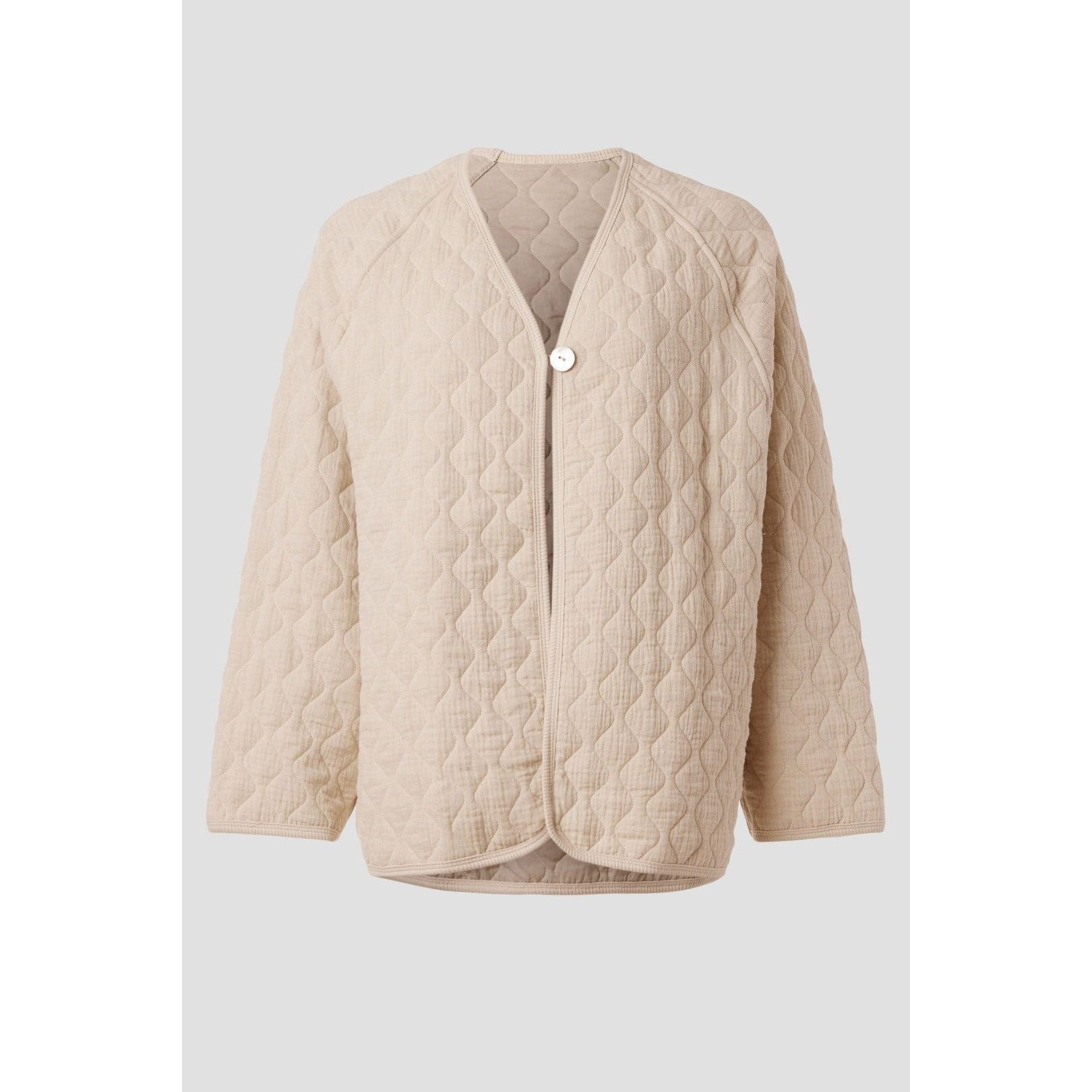 The Donni The Quilted Jacket Creme by Donni is a beige quilted jacket with a wavy pattern, made from organic cotton. It features a relaxed, open-front design with long sleeves and a single button closure against a plain background.