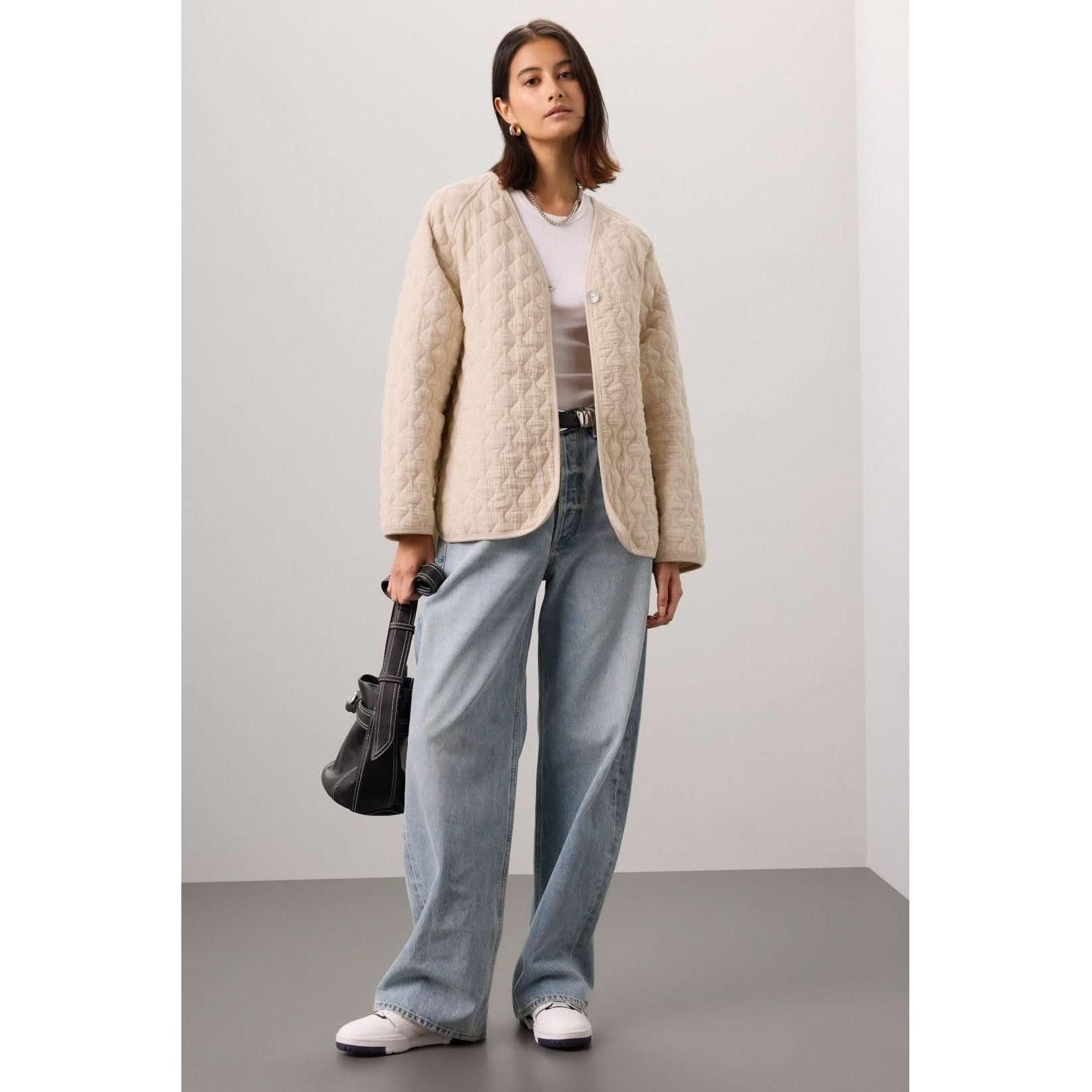 A person poses in the Donni The Quilted Jacket Creme over an organic cotton white top, paired with light blue wide-leg jeans and white sneakers. They hold a black handbag, standing against a plain, light-colored background.