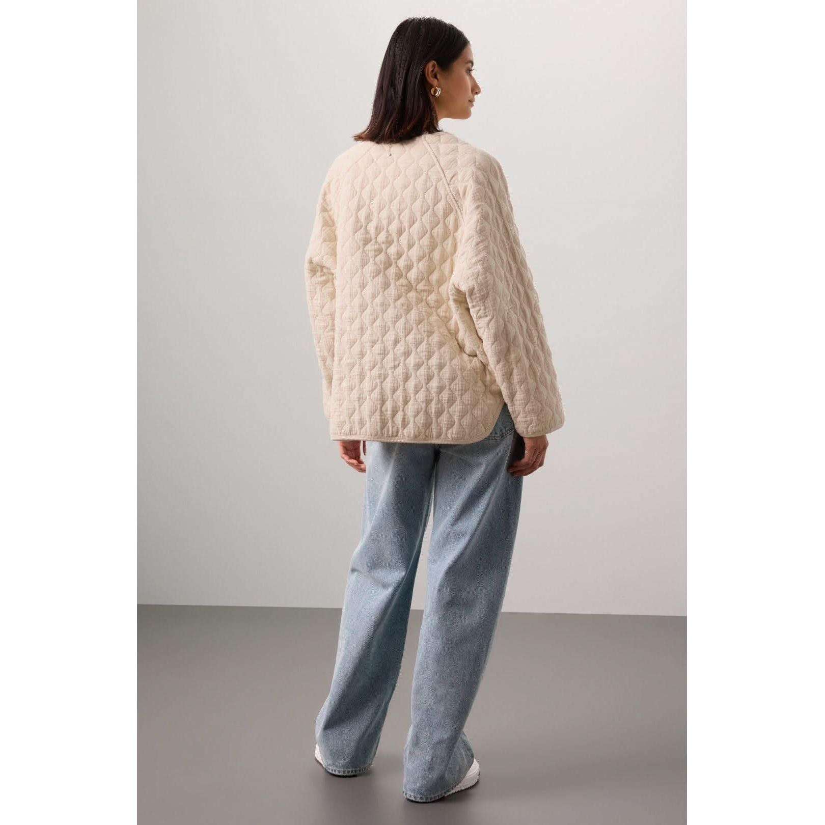 A person with long dark hair stands facing away, wearing the Donni The Quilted Jacket Creme made from organic cotton and light blue jeans. The setting is minimalistic with a gray background and floor.