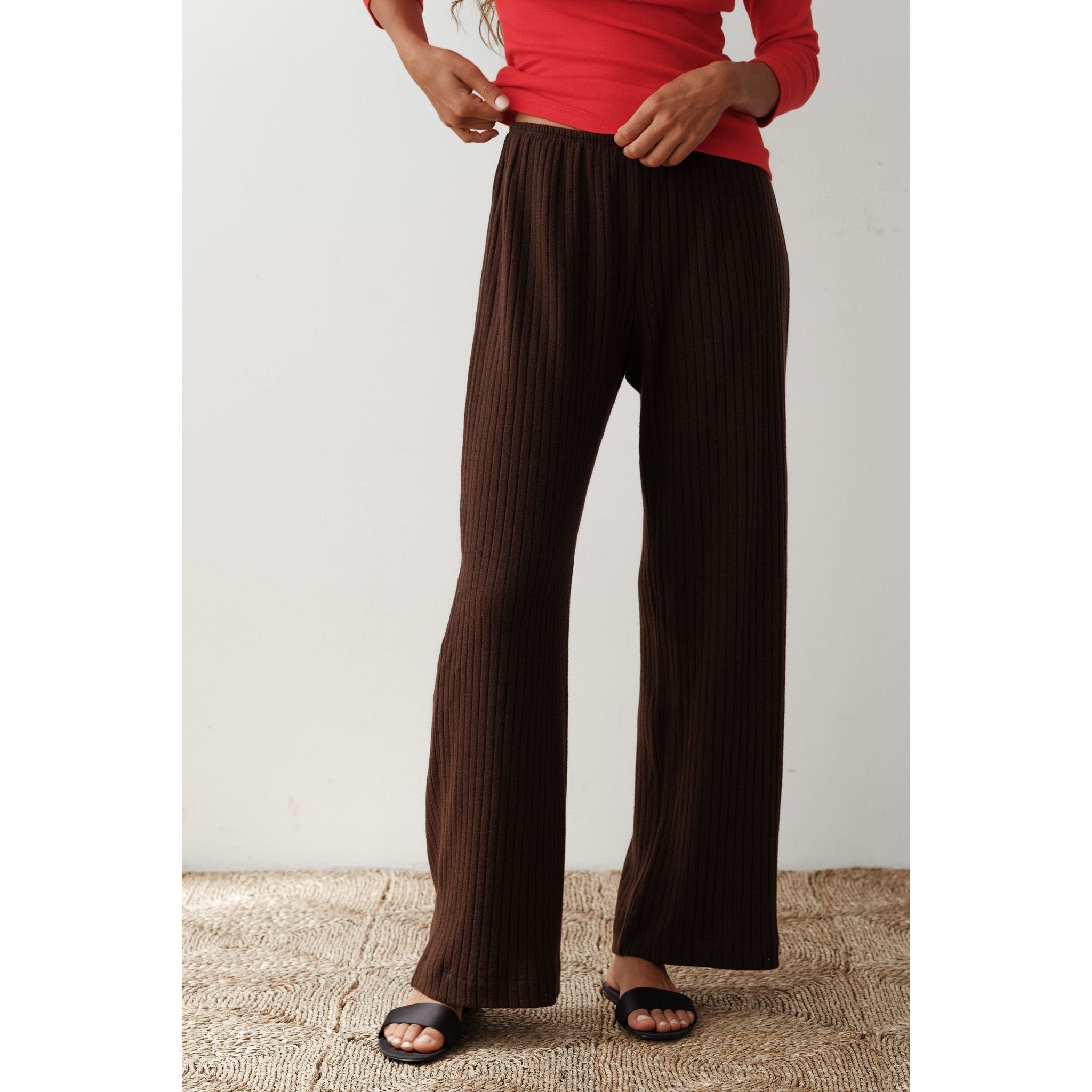 A person stands on a textured rug wearing Donni The Sweater Rib Simple Pant in Espresso, along with a red long-sleeved top and black slide sandals. The photo captures their legs and torso, but not their face.