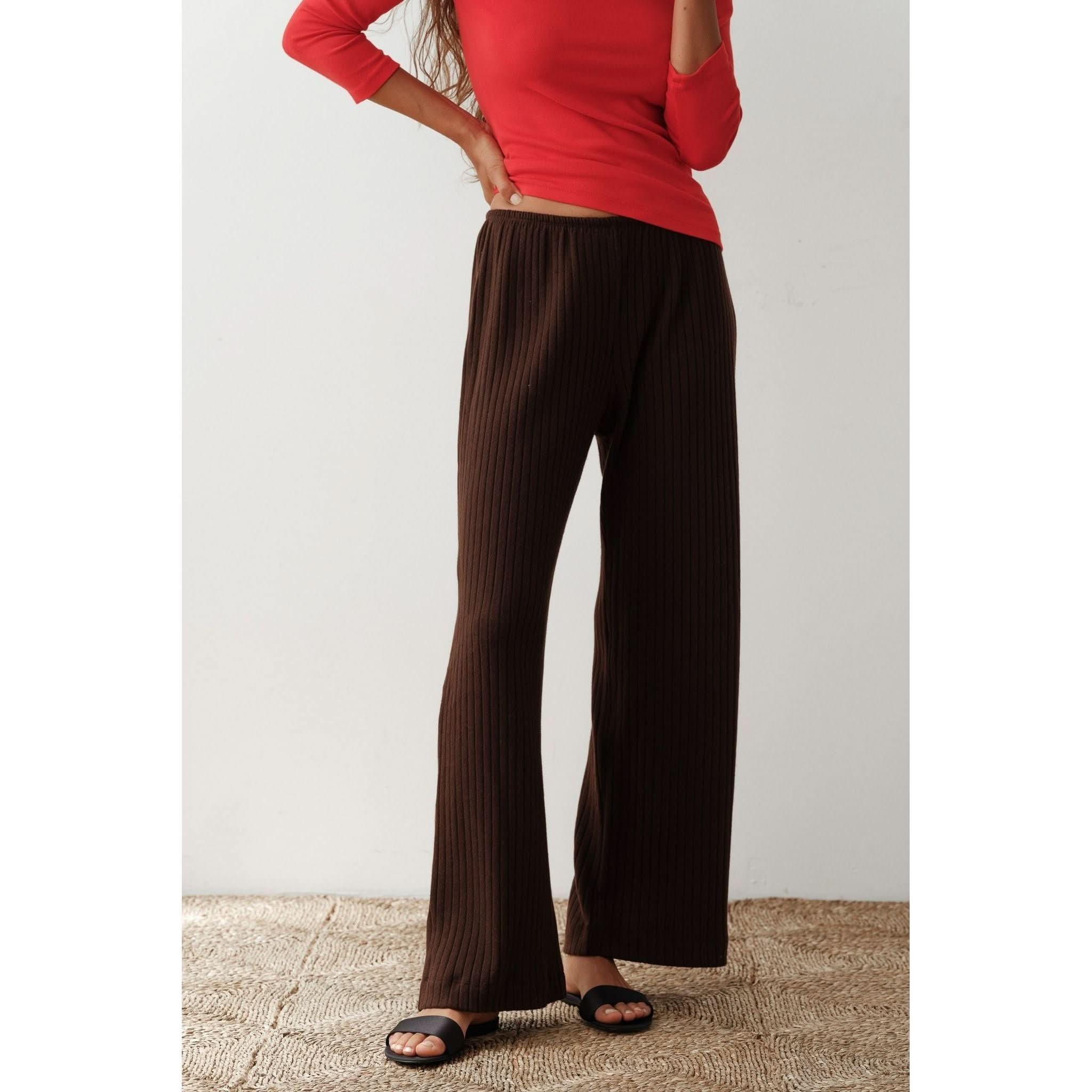 A person wearing the Donni The Sweater Rib Simple Pant in Espresso and a red long-sleeve top made from organic cotton, along with black slide sandals, stands on a textured rug against a plain white background. The person's face is partially out of frame.