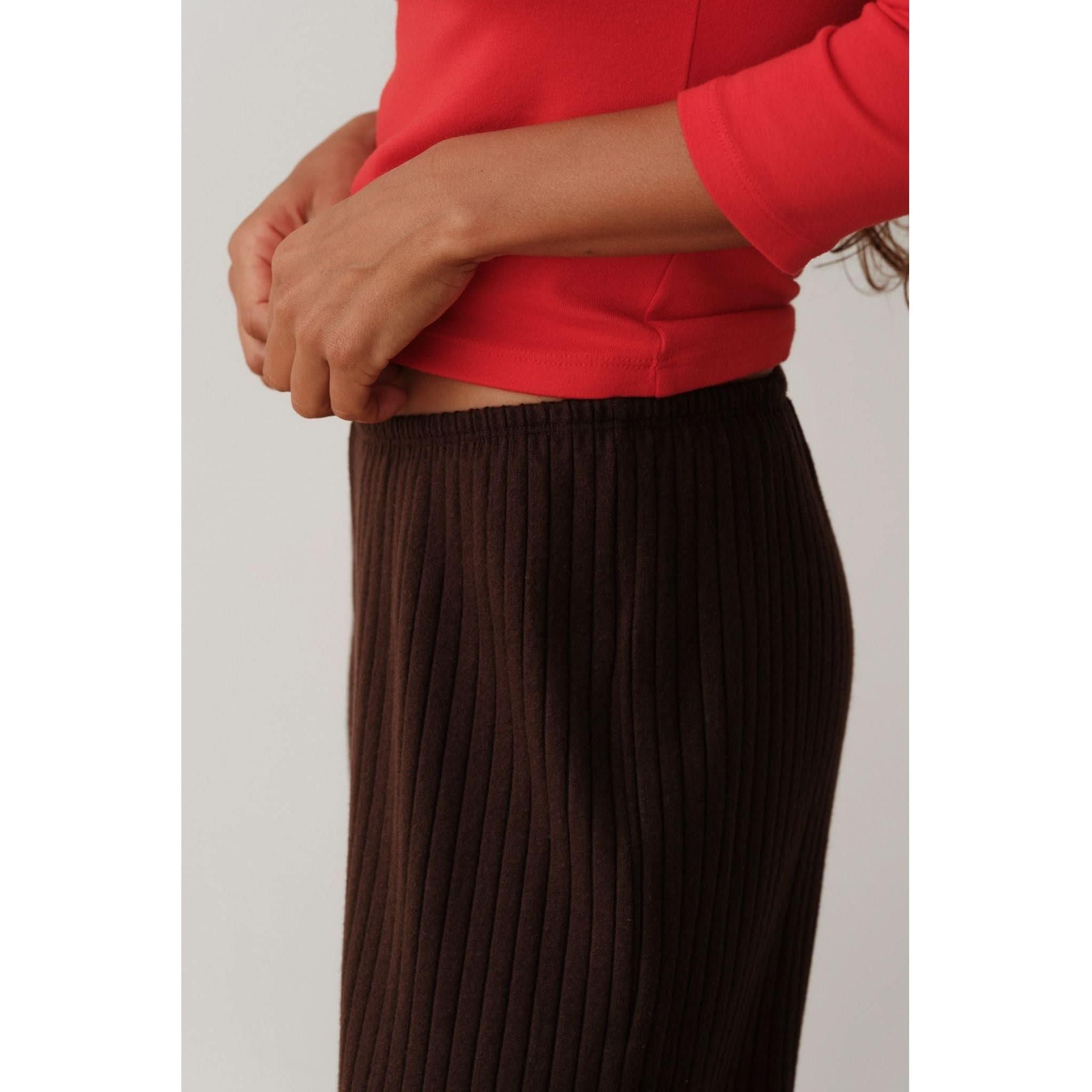 A person is adjusting a pair of espresso-colored pants from Donni, known as The Sweater Rib Simple Pant. These ribbed pants feature an elastic waistband and are worn with a red organic cotton long-sleeve top. The image emphasizes the super-soft texture of the sweater rib fabric against a plain background.