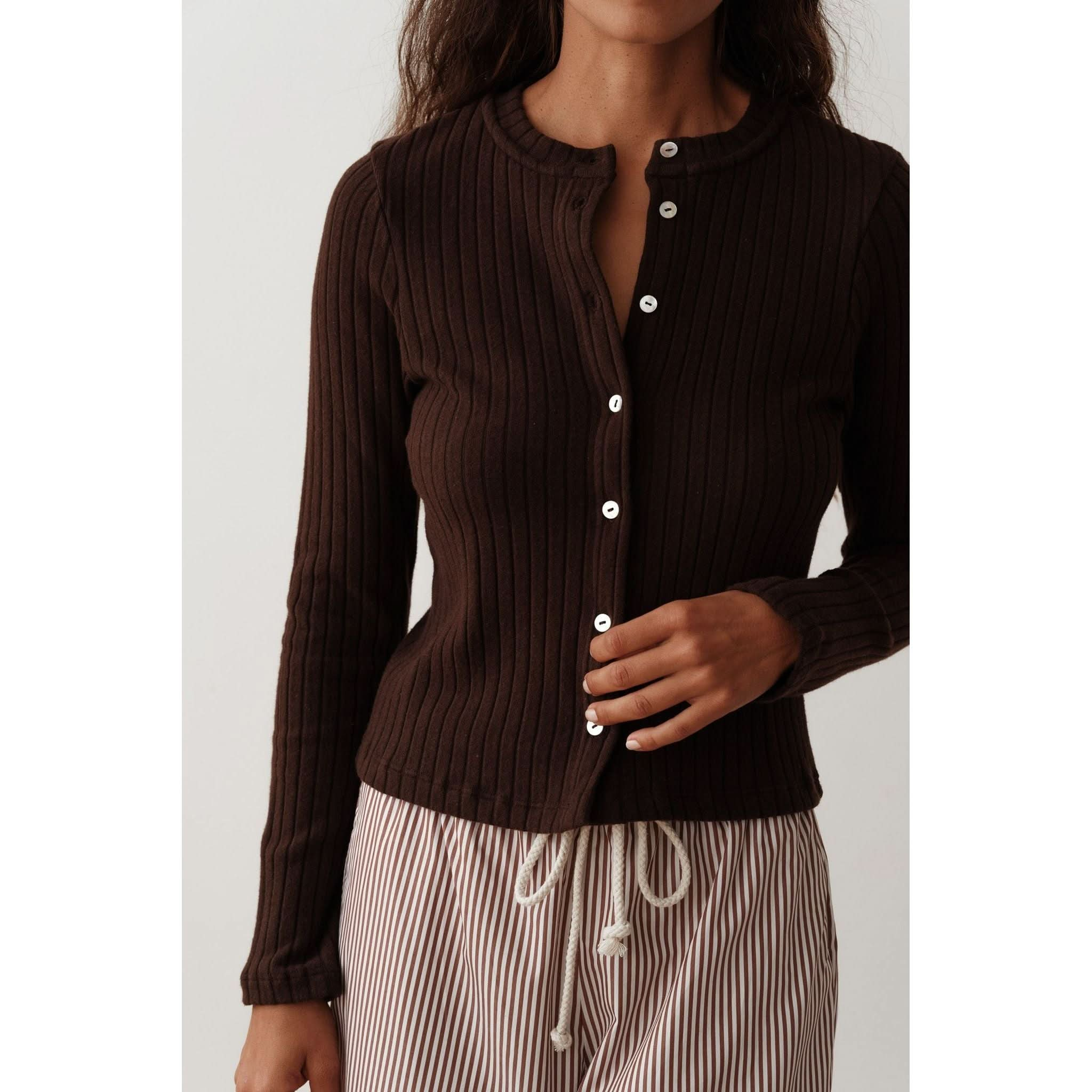 A woman in the Donni The Box Weave Stretch Cardi Jet, a ribbed dark brown cardigan with a crew neck and button placket, is partially buttoning it. She pairs it effortlessly with striped beige and white pants featuring a drawstring against a simple, elegant white background.