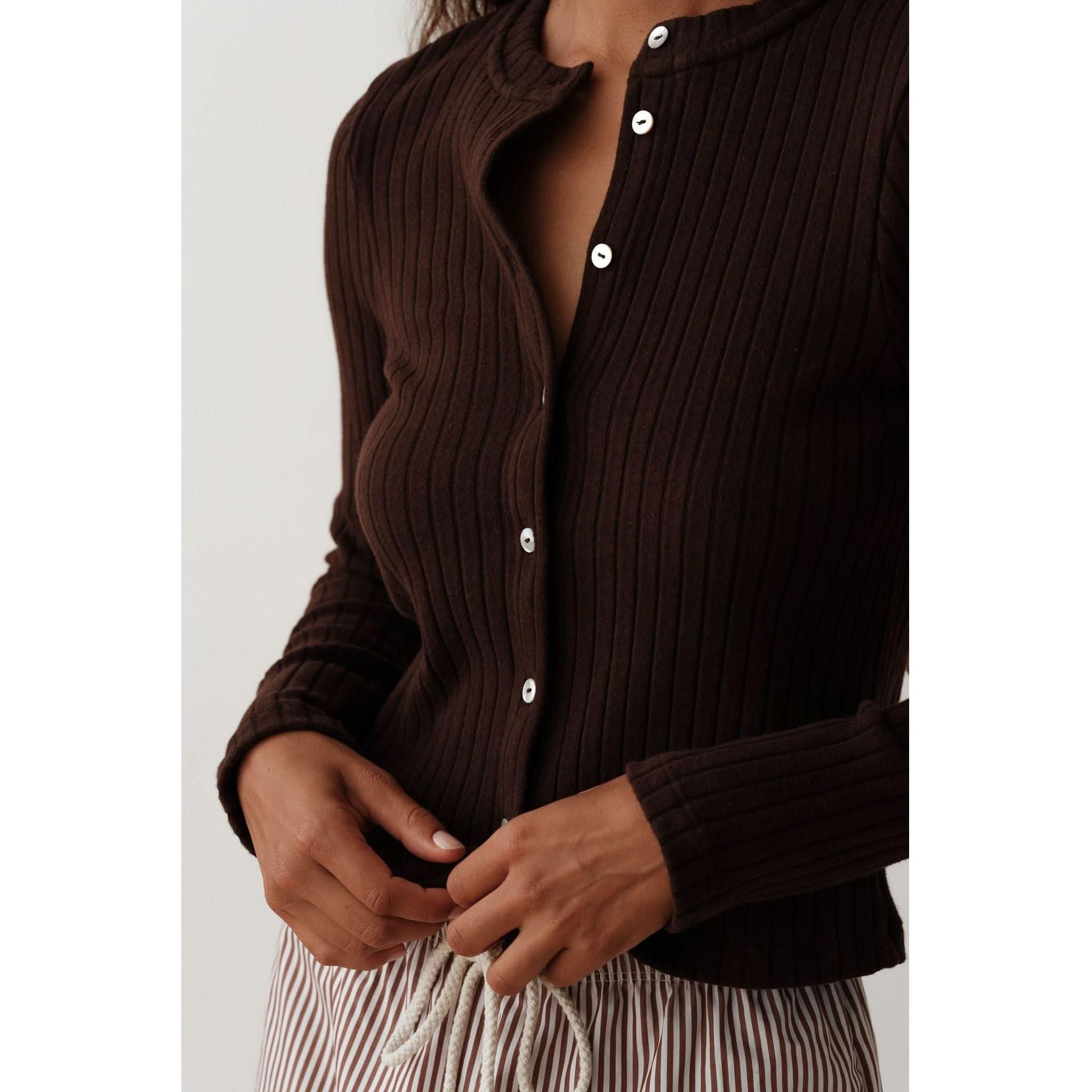 A person wearing the Donni The Sweater Rib Cardi Espresso, a fitted cardigan with ribbed texture and mother of pearl buttons, along with striped pants. The cardigan is partially unbuttoned as the person stands against a plain light background.