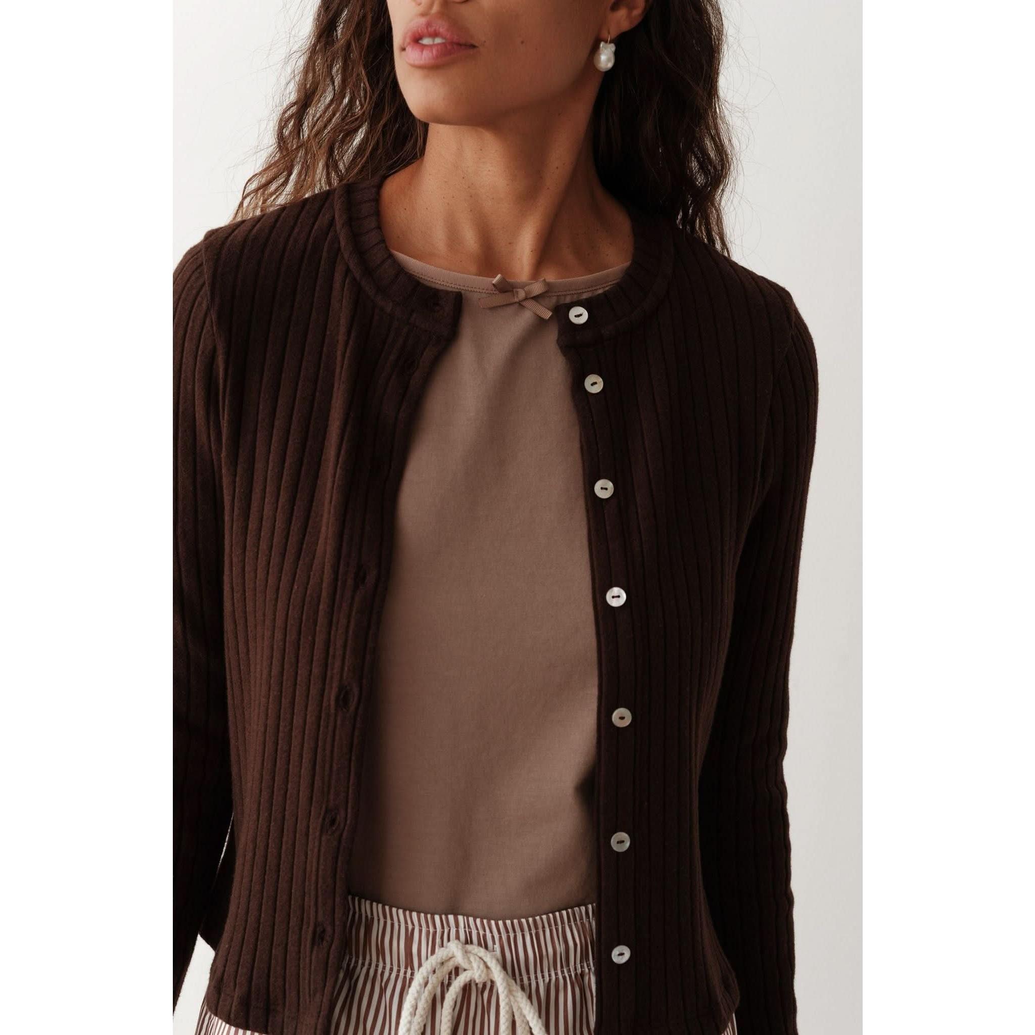 A person wearing the Donni The Sweater Rib Cardi in Espresso, featuring mother of pearl buttons, with a taupe top underneath and accessorized with pearl earrings. Their wavy hair enhances the elegance of their outfit as they pose against a plain background.