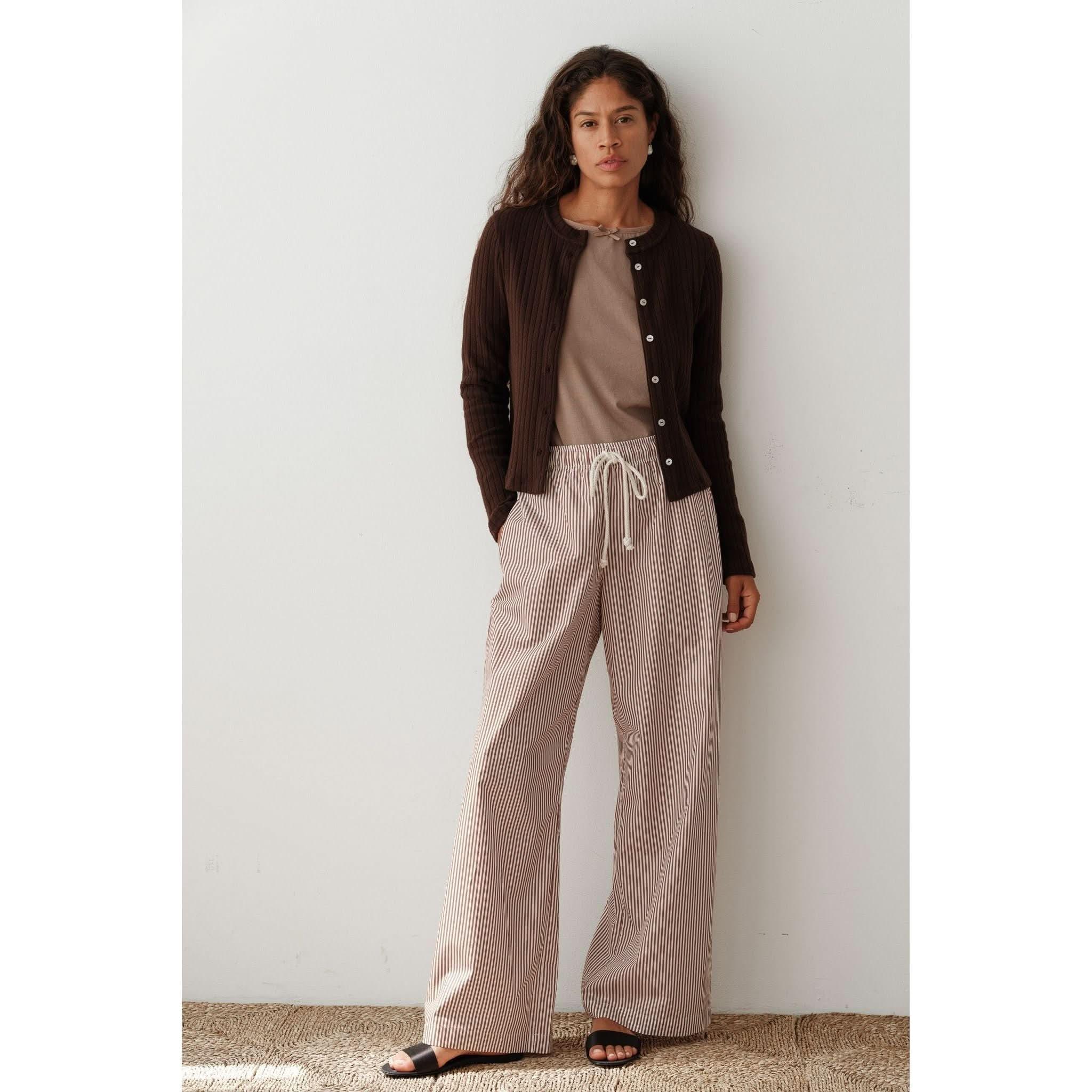 A person with long hair stands against a white wall, wearing the Donni The Sweater Rib Cardi in Espresso, a fitted cardigan adorned with mother of pearl buttons, over a beige top. They pair it with striped wide-leg trousers featuring drawstrings and complete the look with black sandals.