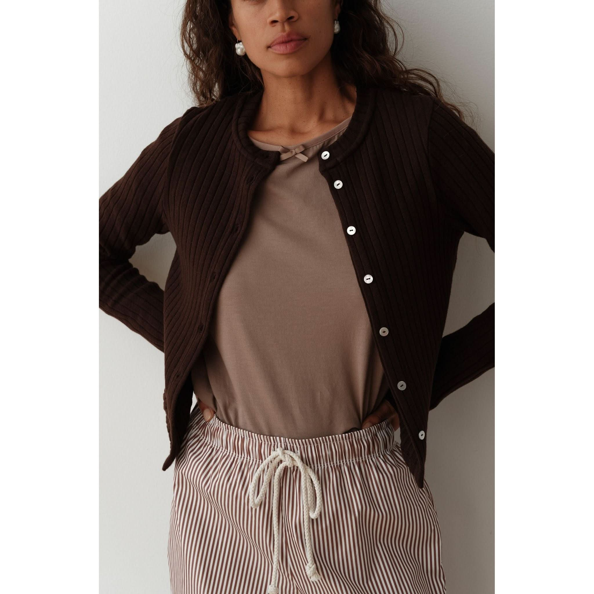 A person stands against a neutral background, wearing the Donni The Box Weave Stretch Cardi Jet, featuring a button placket over a beige top. They are also dressed in striped pants with a drawstring. The person's hair is wavy and loose.