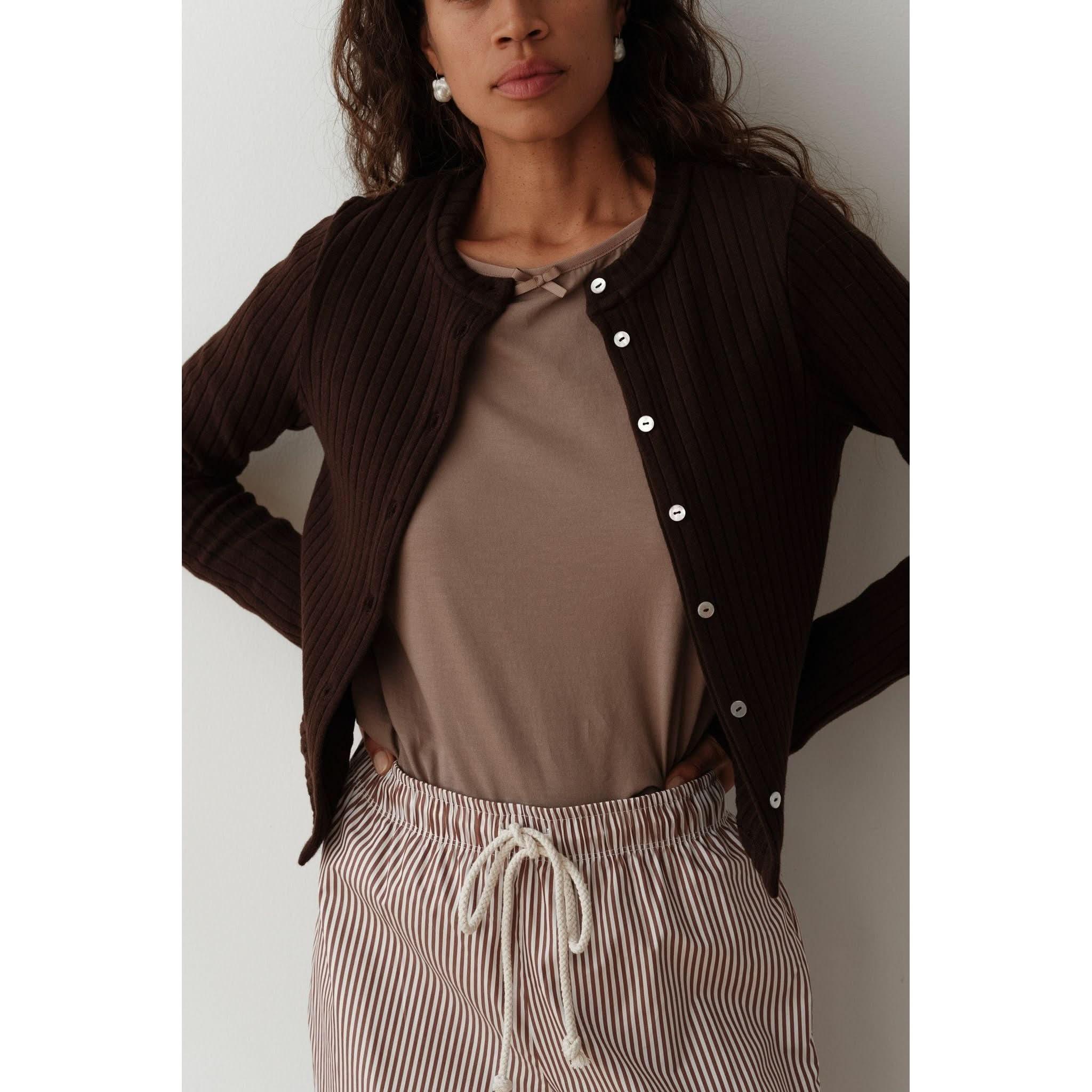 A person is wearing the Donni The Sweater Rib Cardi in Espresso, a fitted dark brown cardigan with mother-of-pearl buttons, layered over a taupe T-shirt. They have paired it with brown and white striped pants featuring a white drawstring, standing against a plain, light-colored background.