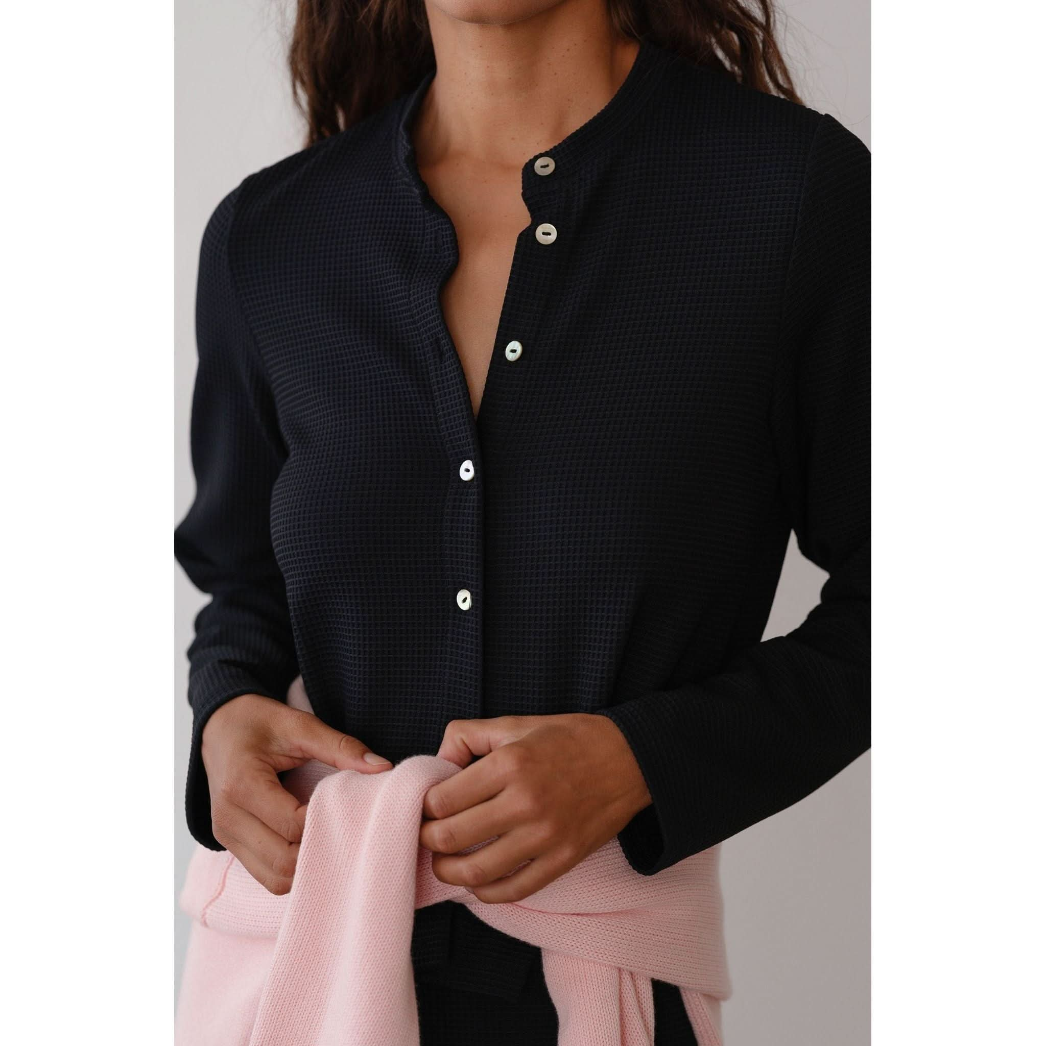 A person wearing a dark, textured button-up shirt with a classic button placket and the Donni The Box Weave Stretch Cardi Jet tied around their waist. The photo highlights the upper body and the cardi, emphasizing its subtle waffle weave texture against a minimal background.