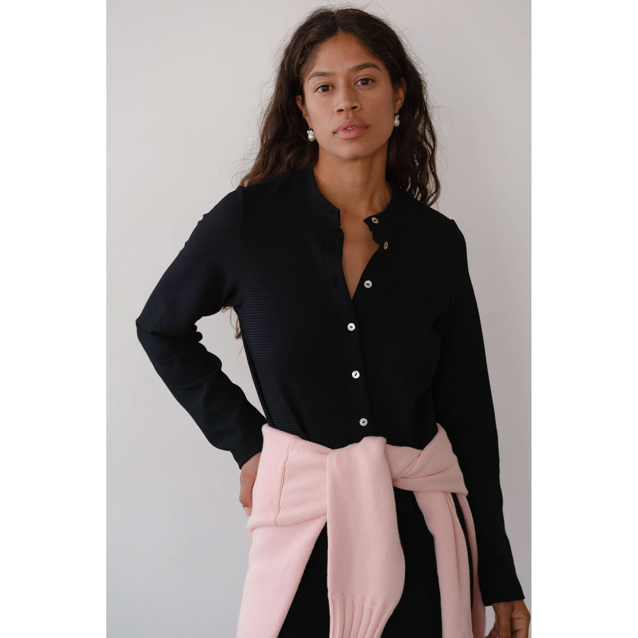 A woman with long, wavy hair is wearing Donni's The Box Weave Stretch Cardi Jet. She has a light pink crew neck sweater tied around her waist and stands against a plain white background, looking into the camera with a neutral expression.