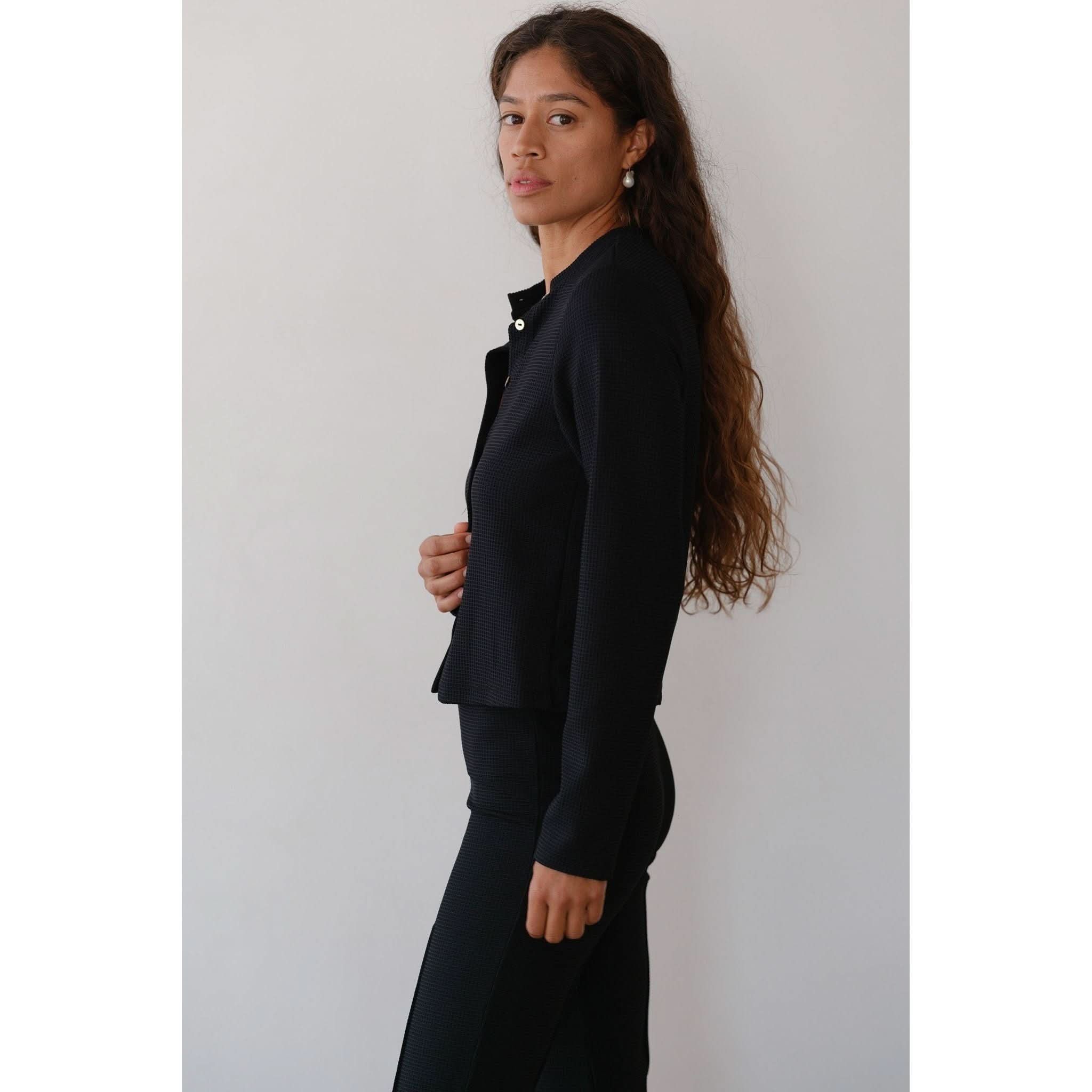 A person with long, wavy hair stands in profile against a light background. They are wearing the stylish Donni The Box Weave Stretch Cardi Jet, a fitted black outfit complete with chic button placket and matching pants, accessorized with pearl earrings.