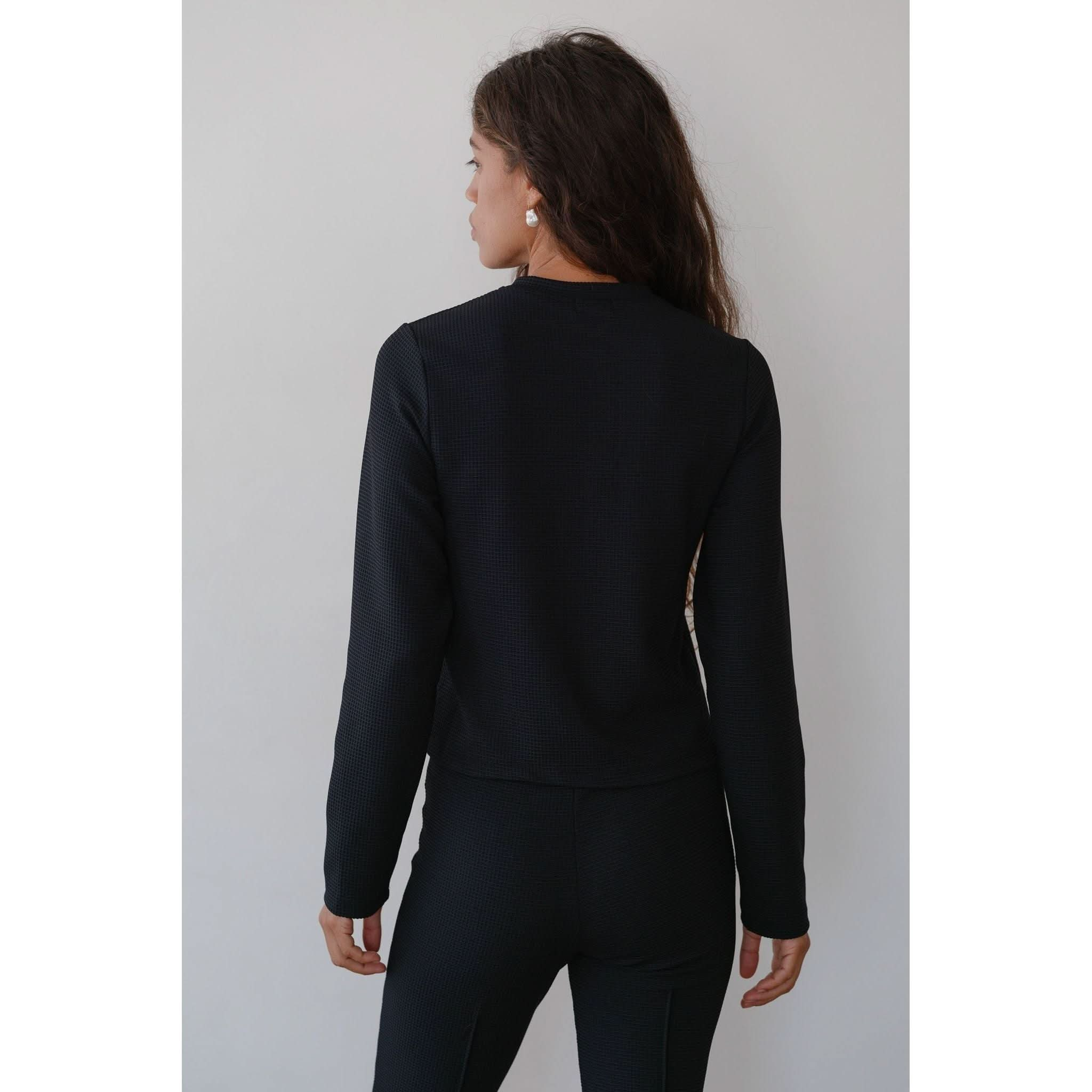 A woman with long brown hair stands with her back to the camera, wearing the Donni The Box Weave Stretch Cardi Jet, a black top featuring a button placket, paired with matching pants against a plain white background. She has a single earring visible on her left ear.