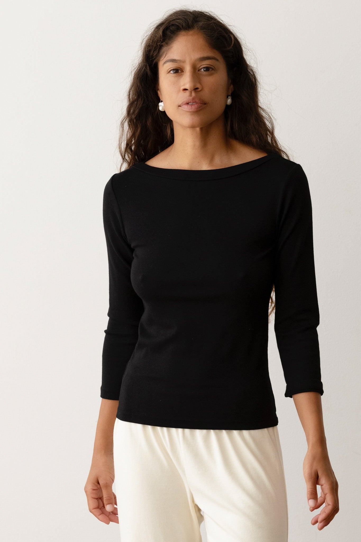 A person with long, wavy hair is wearing the Donni The Baby Rib Boat Neck Black top by Donni, which features 3/4 length sleeves. They have paired it with light-colored pants and are standing against a plain light-colored background.