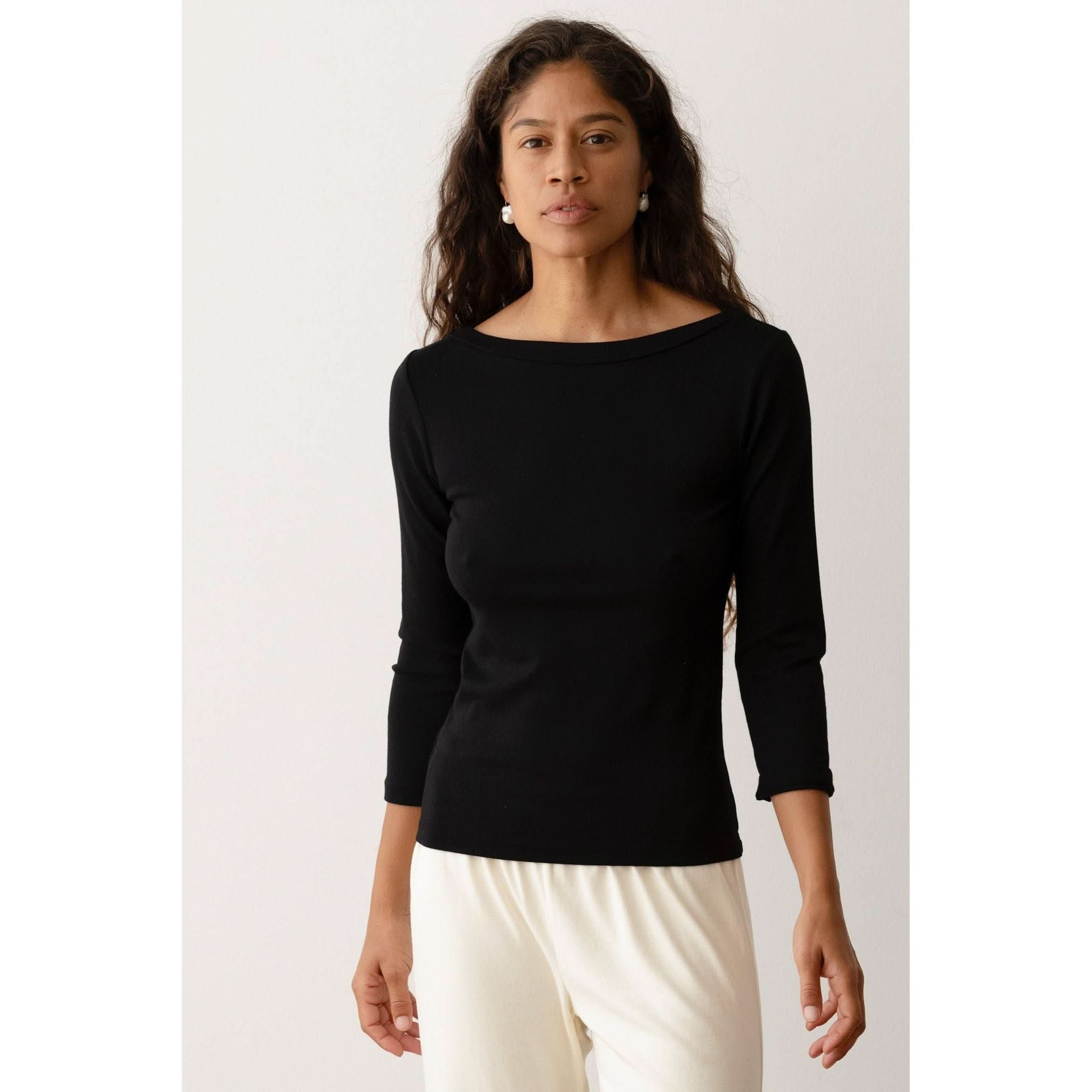 A person with long, wavy hair is wearing the Donni The Baby Rib Boat Neck Black top by Donni, which features 3/4 length sleeves. They have paired it with light-colored pants and are standing against a plain light-colored background.