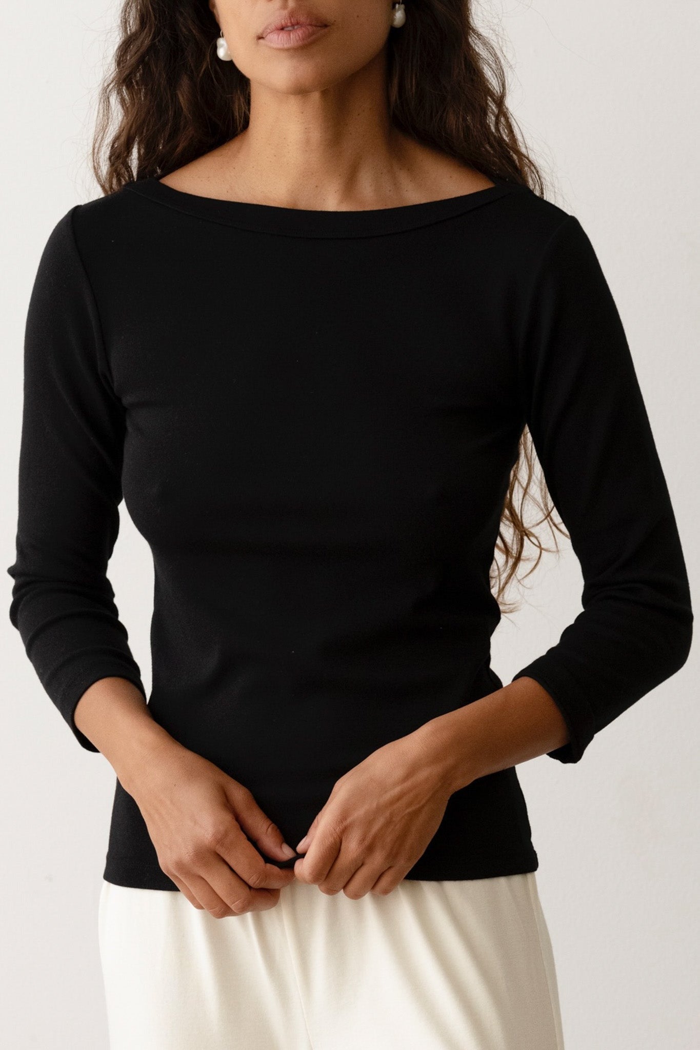 A person with long hair wearing the Donni The Baby Rib Boat Neck Black, a fitted black top made from Baby Rib fabric, and a light-colored skirt stands against a plain background. The boatneck top has 3/4 length sleeves, and the person is seen from the waist up, with their hands clasped in front of them.