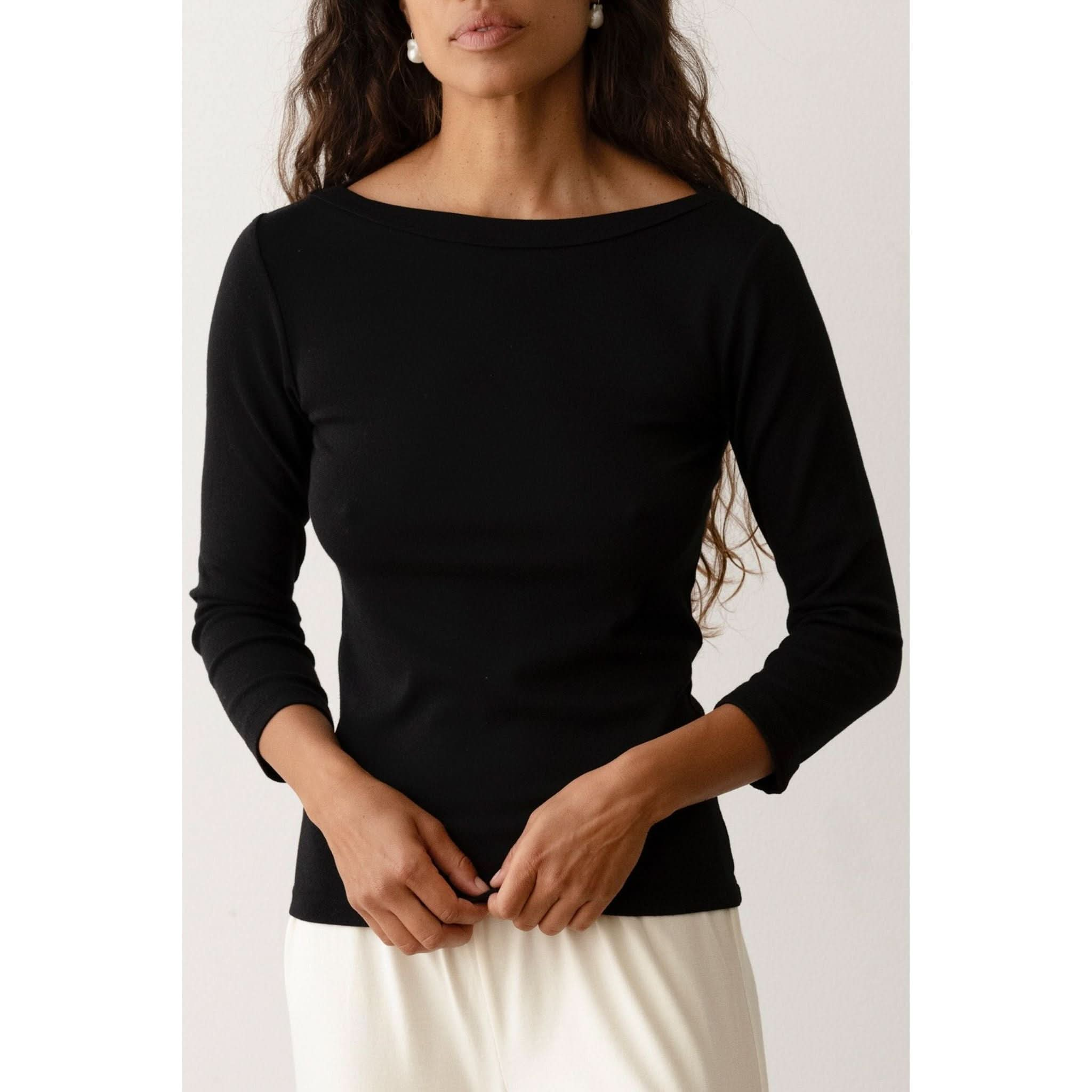 A person with long hair wearing the Donni The Baby Rib Boat Neck Black, a fitted black top made from Baby Rib fabric, and a light-colored skirt stands against a plain background. The boatneck top has 3/4 length sleeves, and the person is seen from the waist up, with their hands clasped in front of them.