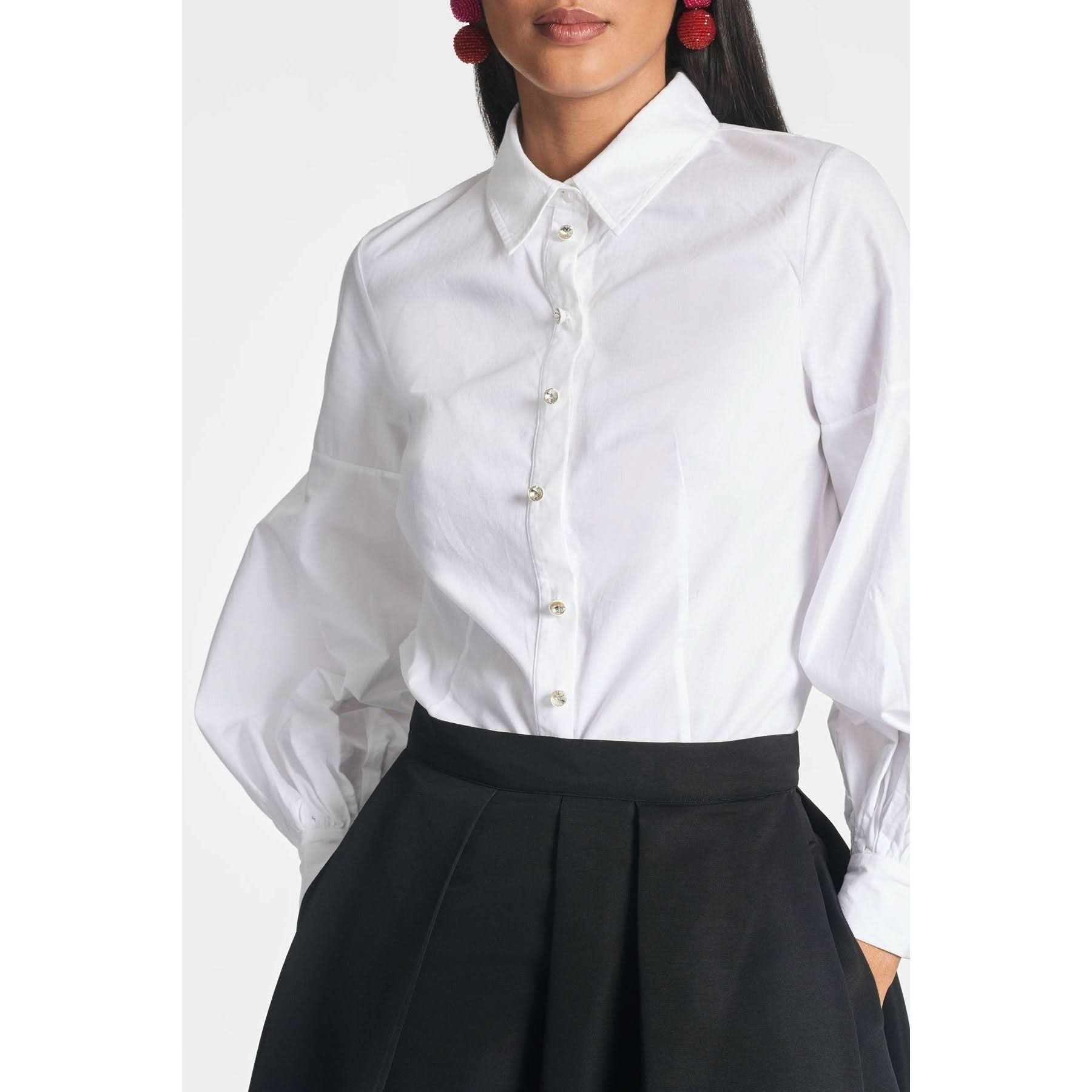 A person wearing a white Sachin & Babi Elizabeth Top with long, slightly puffed balloon sleeves and a black high-waisted skirt. The blouse has a classic collar and is buttoned up, featuring crystal button embellishments. They are also wearing large red earrings. The background is plain and white.