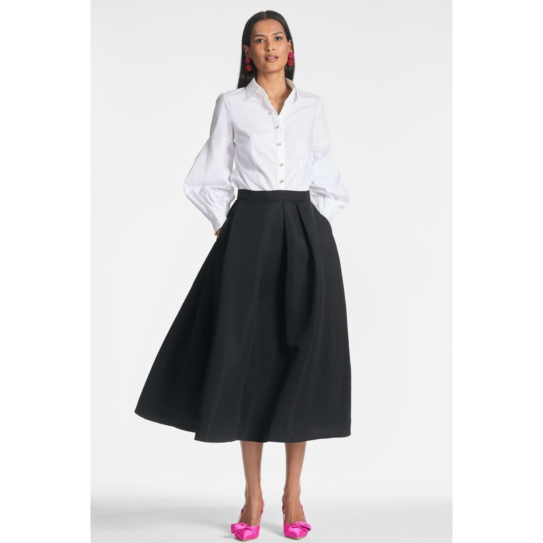 A person stands against a plain background, wearing a Sachin & Babi Elizabeth Top with crystal button embellishments, paired with a high-waisted black midi skirt and bright pink high heels. They have long dark hair and are accessorizing with small, dangling earrings.