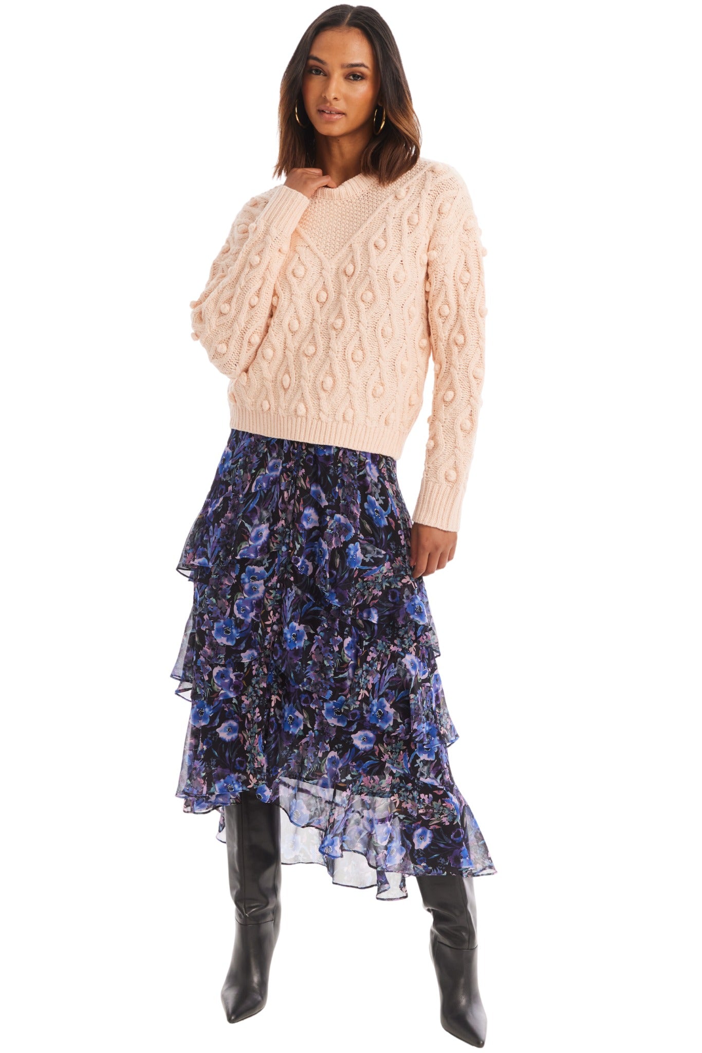 A person stands against a white background, wearing an Allison NY Esme Sweater from the brand Allison, adorned with a swirling cable knit pattern. They pair it with a layered, ruffled skirt showcasing a dark blue and purple floral design. Their ensemble is completed with long, dark hair cascading down and black knee-high boots.