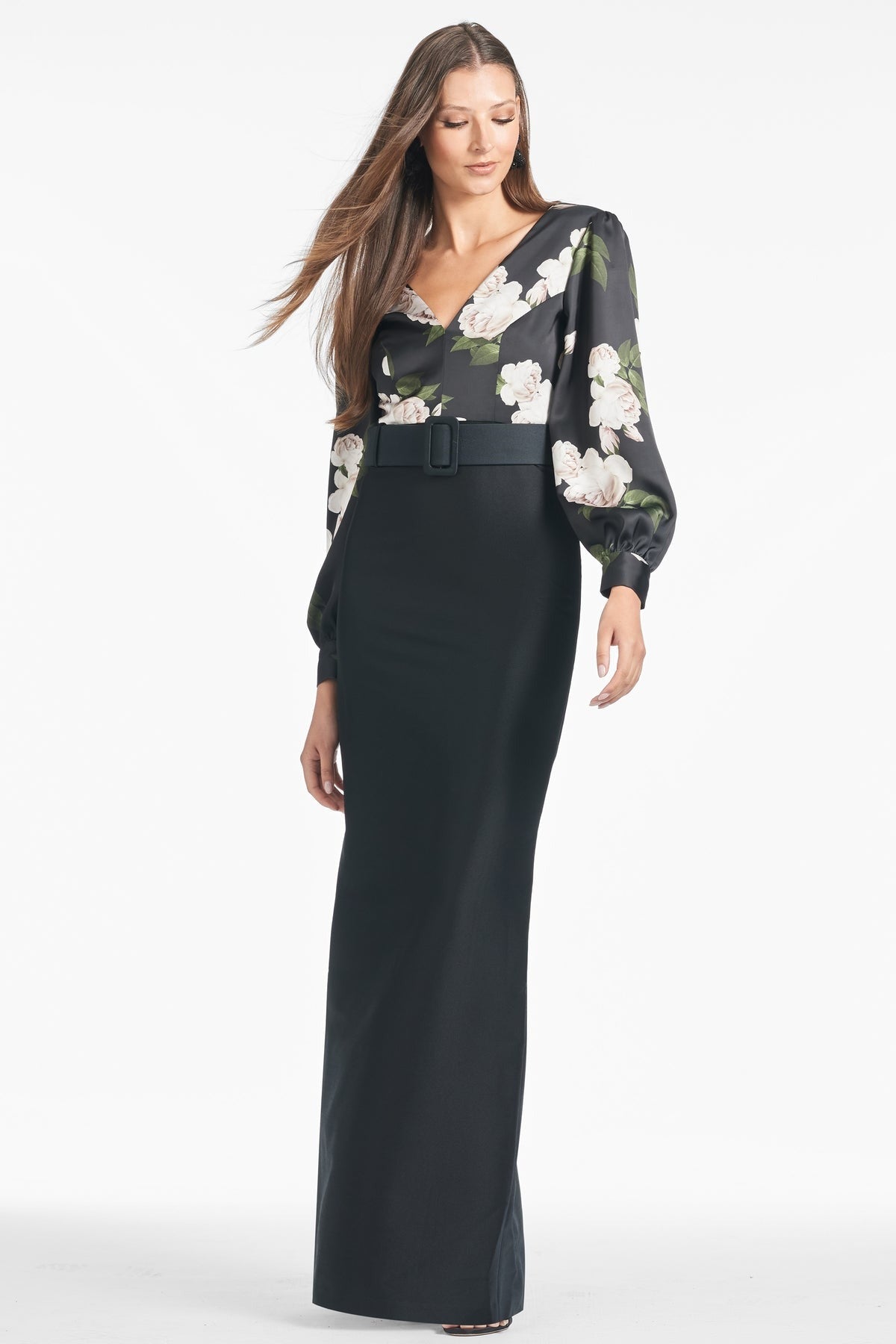 A woman with long hair wears a stylish, full-length Sachin & Babi Schylor Gown in Obsideon Blooms Black, featuring a floral pattern on the upper portion and sleeves and a wide belt at the waist. She stands in a neutral pose against a plain white background.