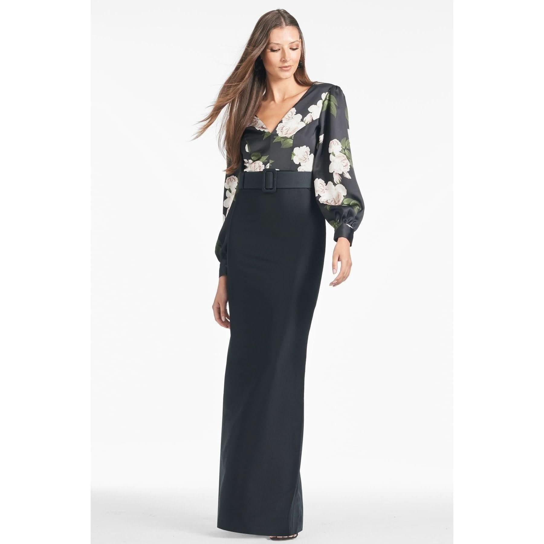 A woman with long hair wears a stylish, full-length Sachin & Babi Schylor Gown in Obsideon Blooms Black, featuring a floral pattern on the upper portion and sleeves and a wide belt at the waist. She stands in a neutral pose against a plain white background.