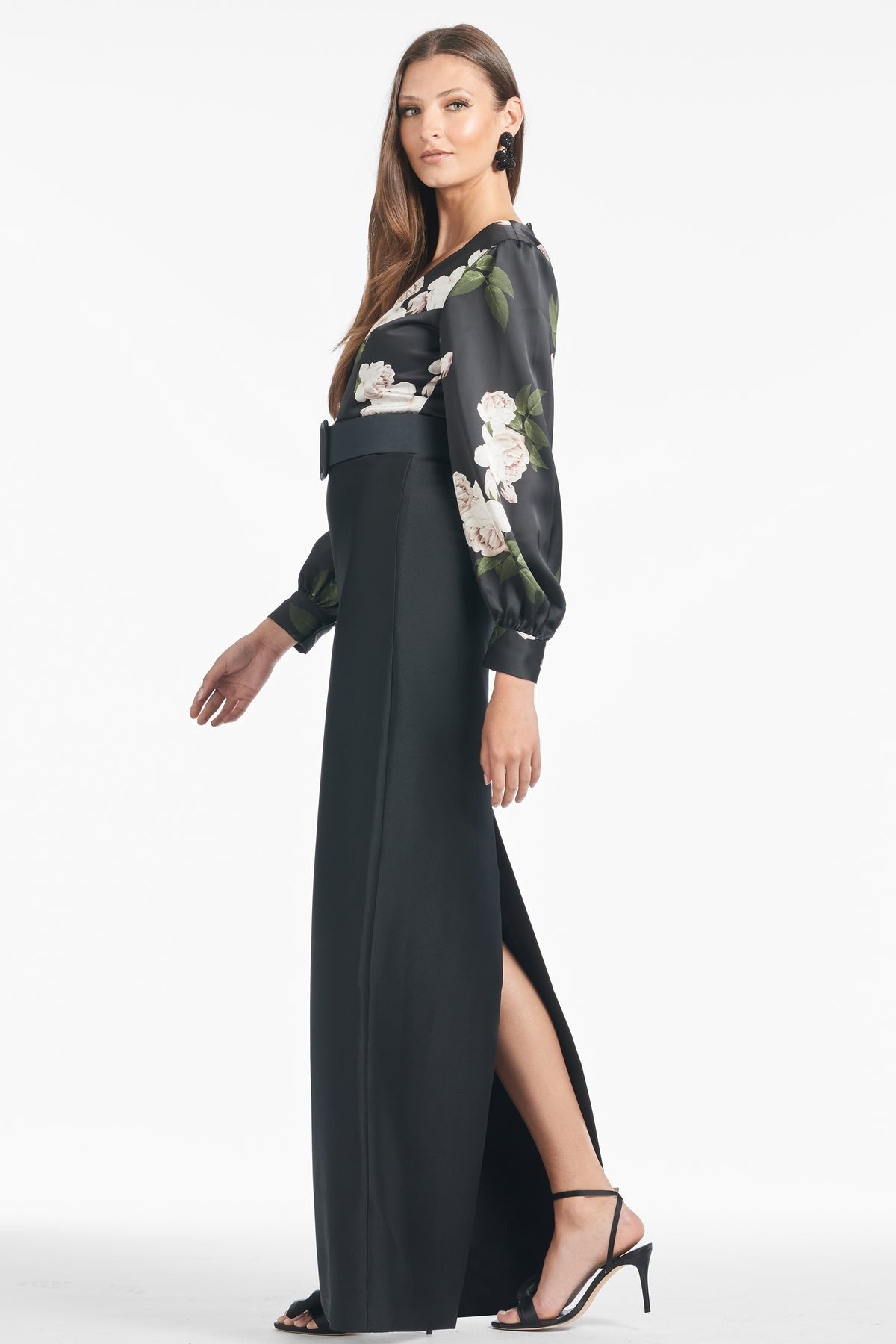 A woman stands in profile, draped in an elegant Sachin & Babi Schylor Gown - Obsideon Blooms Black. The long black skirt with a high slit reveals one leg, while her blouse flaunts the intricate Obsideon Blooms Black print with sheer sleeves. She completes the look with black high-heeled sandals and straight hair, set against a plain white background.