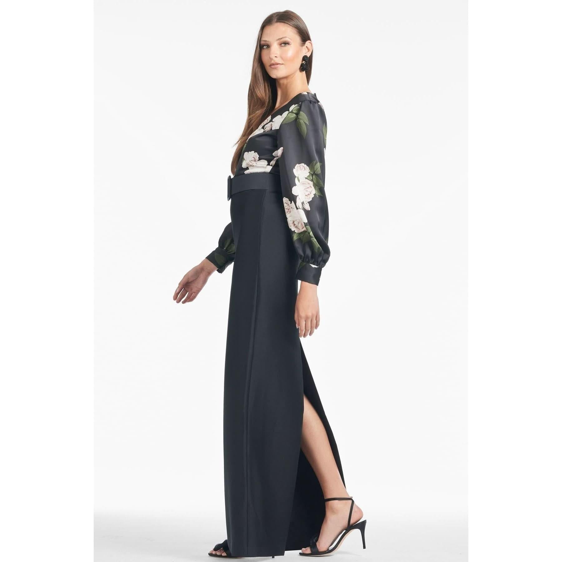 A woman stands in profile, draped in an elegant Sachin & Babi Schylor Gown - Obsideon Blooms Black. The long black skirt with a high slit reveals one leg, while her blouse flaunts the intricate Obsideon Blooms Black print with sheer sleeves. She completes the look with black high-heeled sandals and straight hair, set against a plain white background.