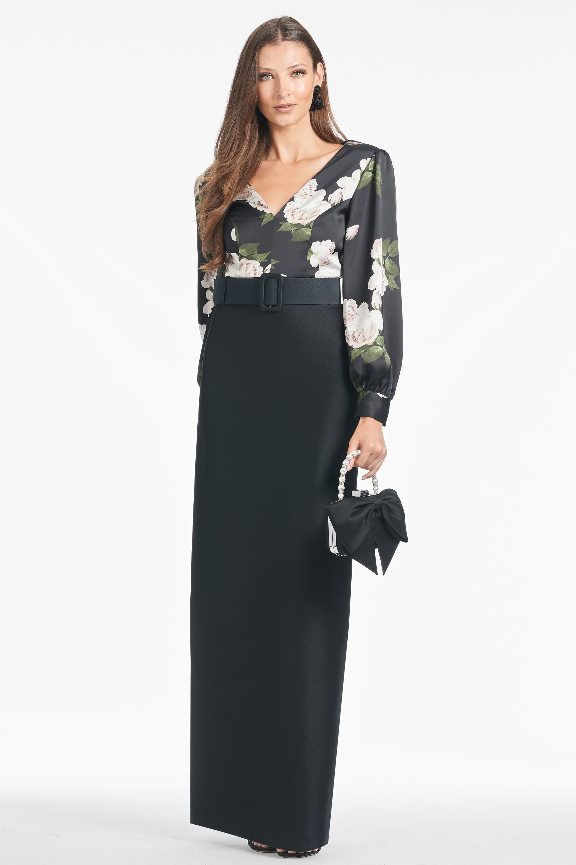 A woman poses in a sophisticated Sachin & Babi Schylor Gown, which features the Obsideon Blooms Black print on the top portion and long sleeves. She holds a black handbag with a bow detail in her right hand. With long, straight hair and small hoop earrings, she stands against a plain white background.