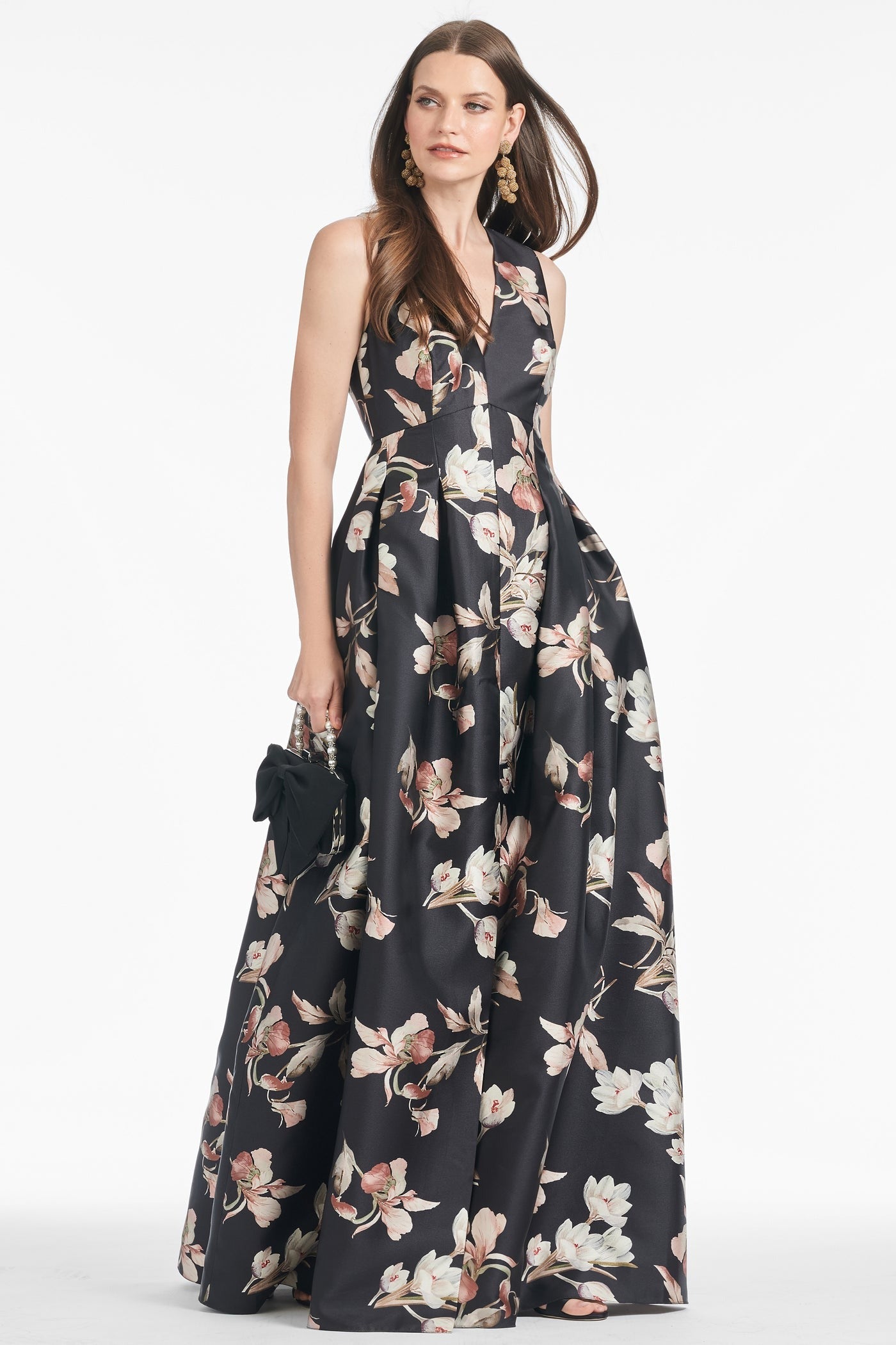 A woman stands wearing the Sachin & Babi Brooke Gown - Tonal Petal, a sleeveless, V-neck dress adorned with large white and pink floral prints. The floor-length black gown features elegant box pleats and discreet side pockets. She holds a small black clutch in her left hand and wears long, dangling earrings. Her straight hair is styled down as she gazes off-camera.